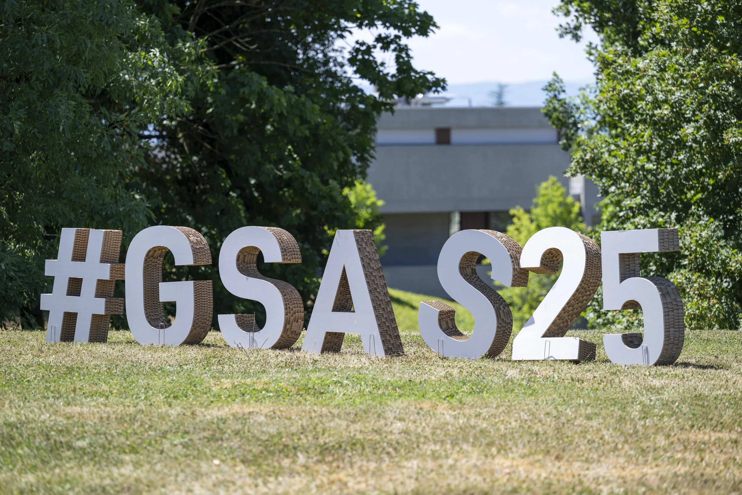 Signes en lettres blanches et brunes indiquant '#GSA S25' dans un espace extérieur herbeux, entouré d'arbres et de bâtiments en arrière-plan.