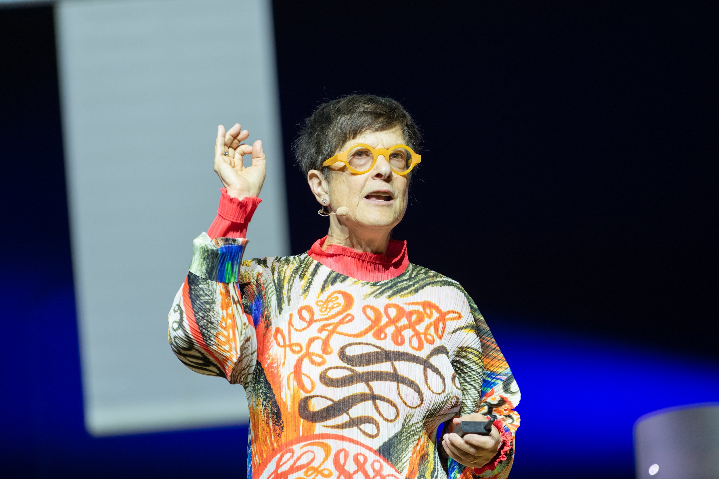 Une femme portant des lunettes jaunes et un pull coloré donne une présentation avec un micro attaché à l'oreille, tenant un pointeur ou un clic. Elle semble parler lors d'une conférence ou d'un événement public.