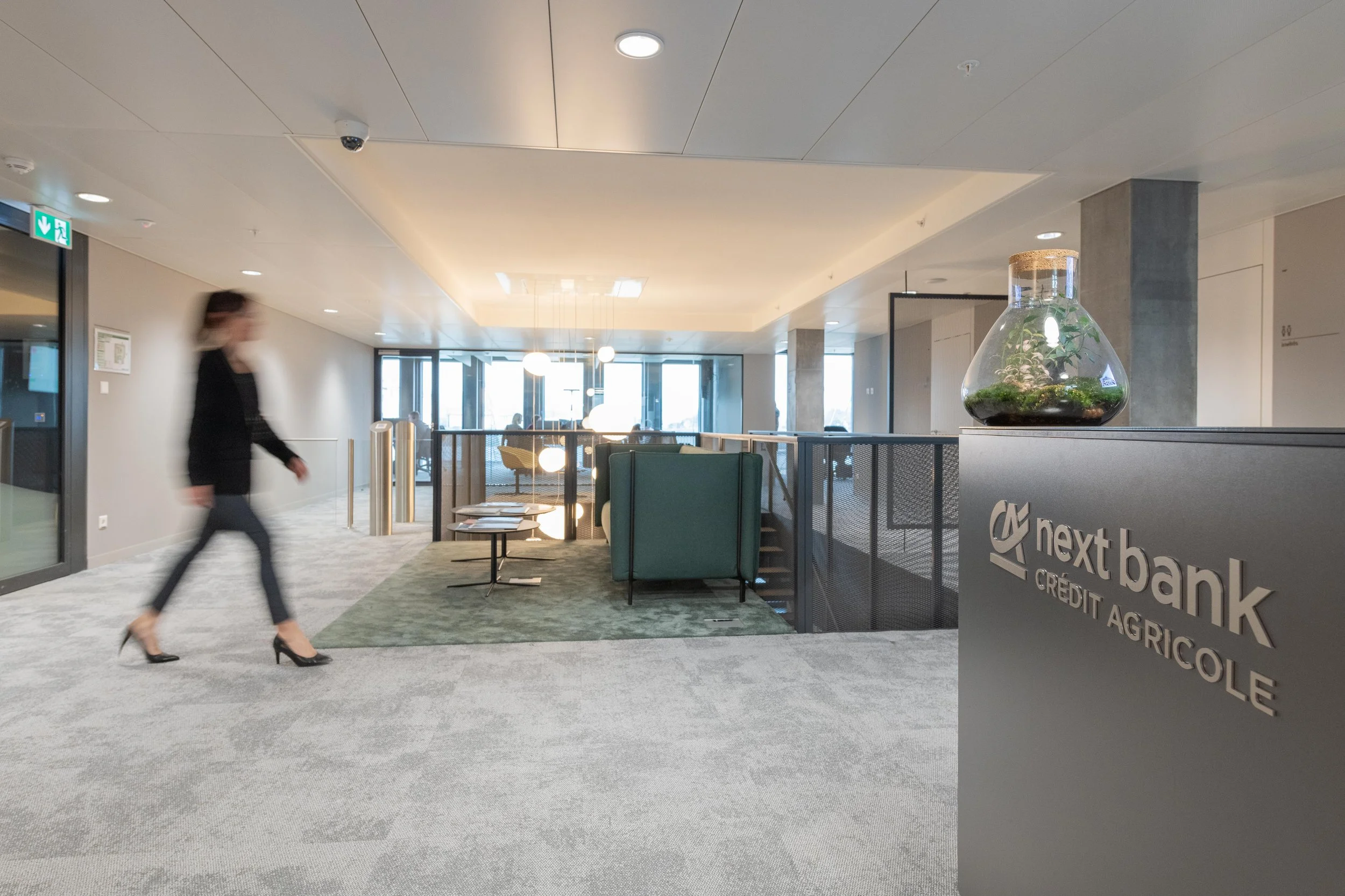 Intérieur d'une banque agricole avec une personne floue passant à gauche, une zone d'accueil avec des fauteuils et une petite table, et un décor moderne avec un aquarium en forme de ballon sur un comptoir.