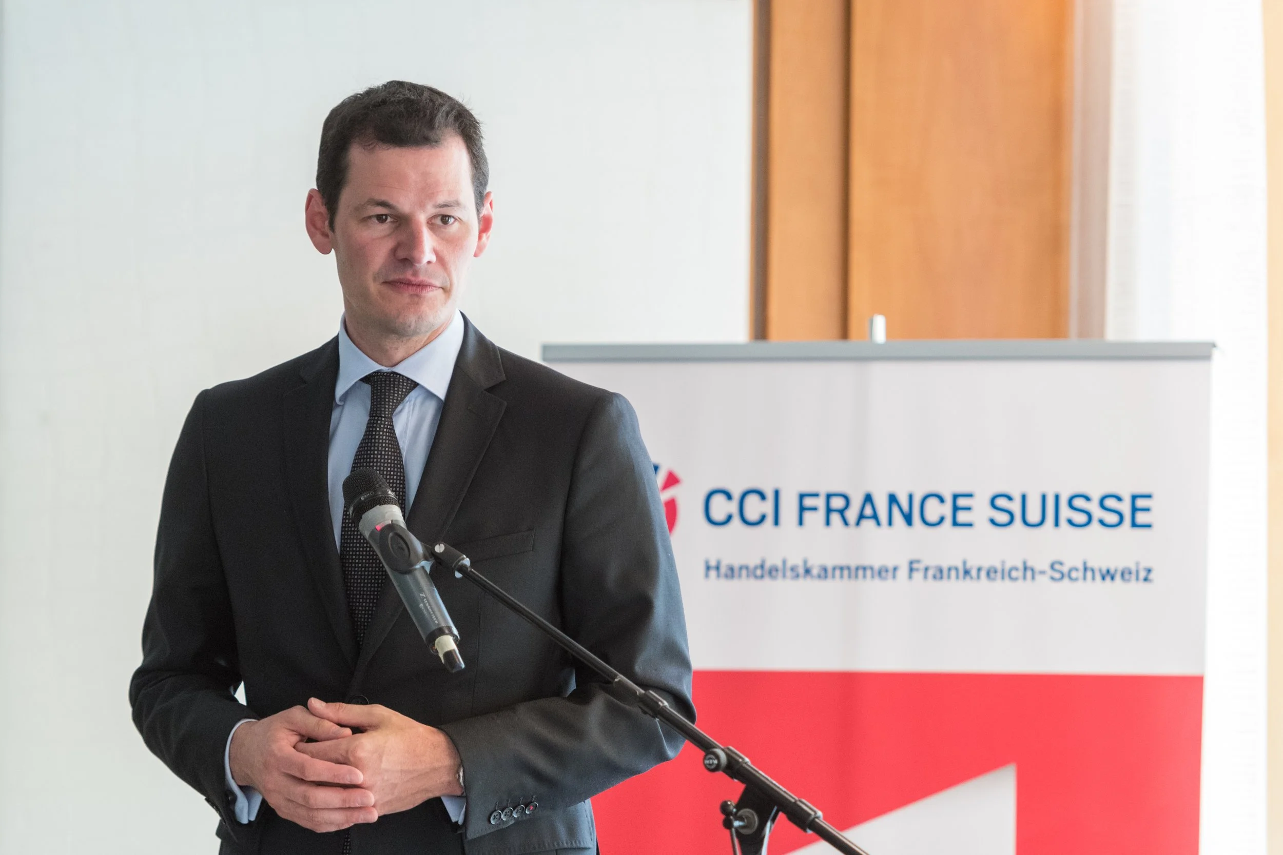Un homme en costume noir parle devant un micro, avec un panneau indiquant 'CCI France Suisse' en arrière-plan.