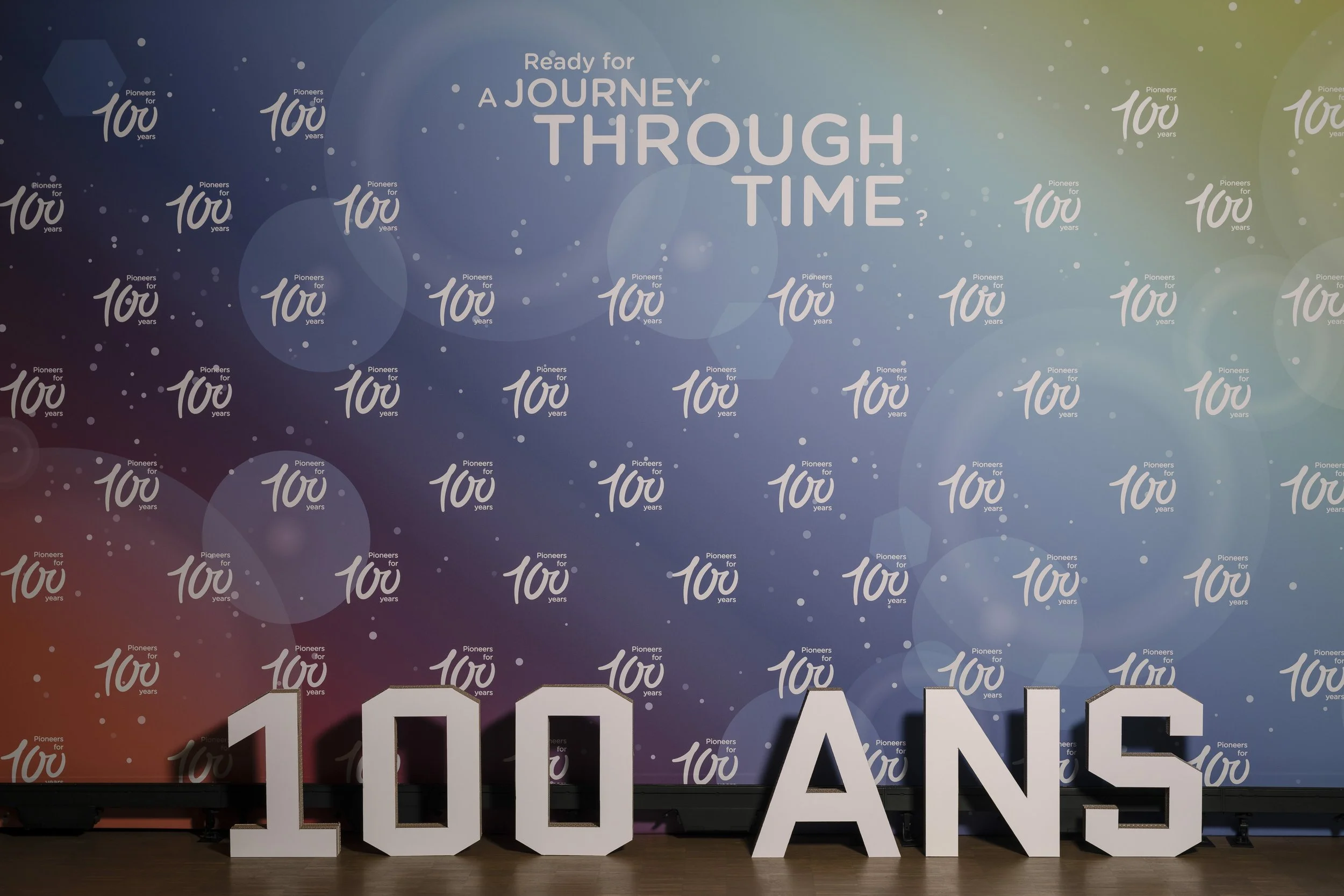 Décoration pour le 100e anniversaire avec des lettres en volume indiquant « 100 ANS » devant un fond festif avec le texte « Ready for a journey through time? »