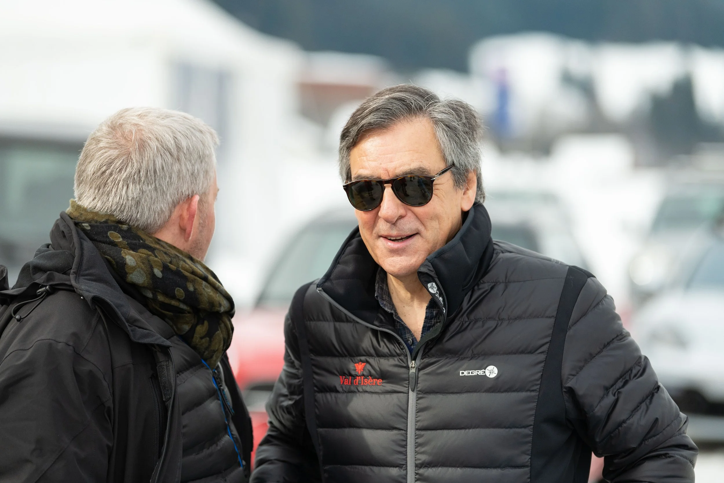 Deux hommes discutent en plein air, portant des vestes noires, avec un homme portant des lunettes de soleil. En arrière-plan, des voitures stationnées dans un parking.