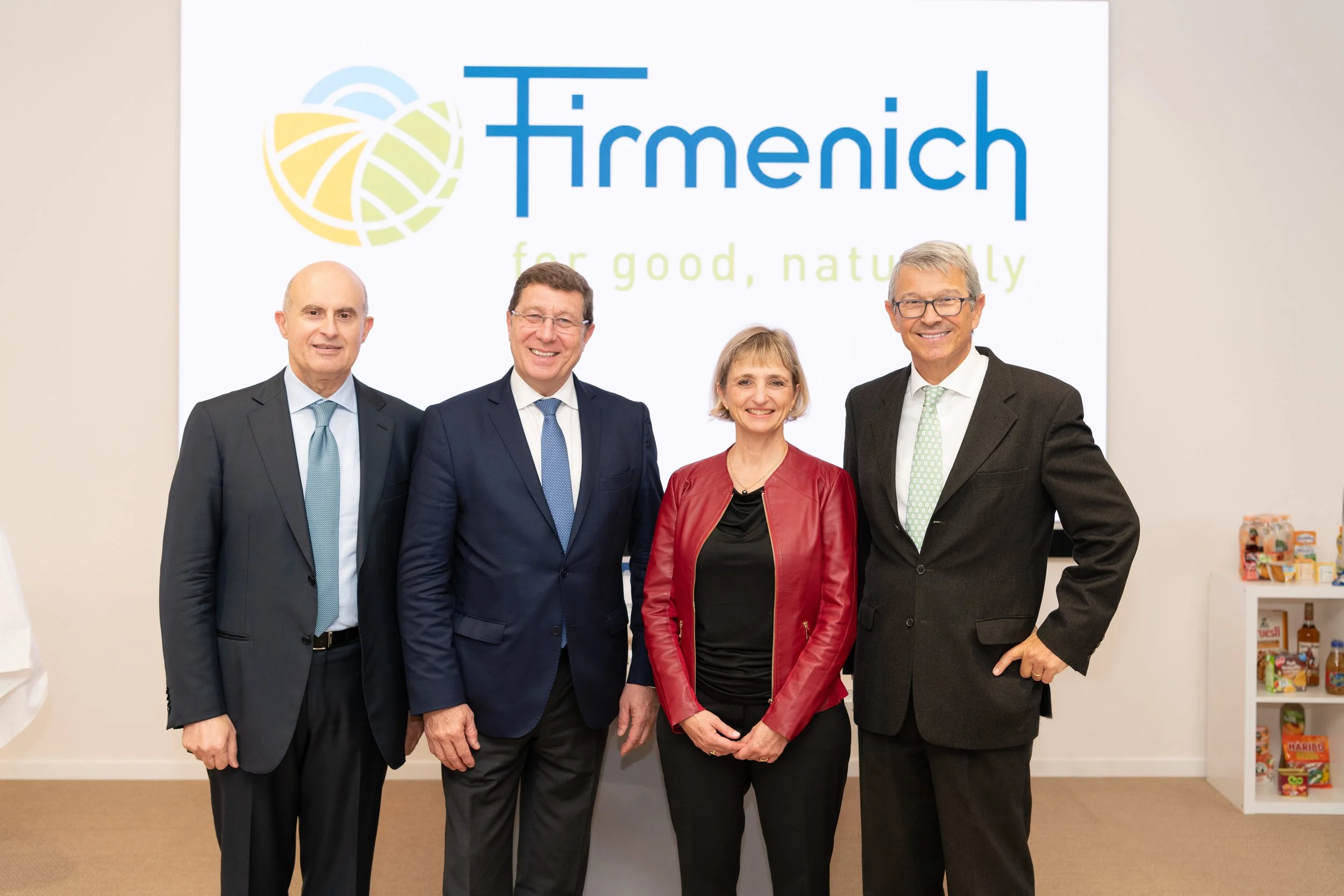 Quatre personnes dans un bureau posant pour une photo avec un logo 'Firmennich' en arrière-plan, slogan 'for good, naturally', vêtements professionnels, homme et femme en costume, femme en veste rouge, meuble avec des snacks à droite.