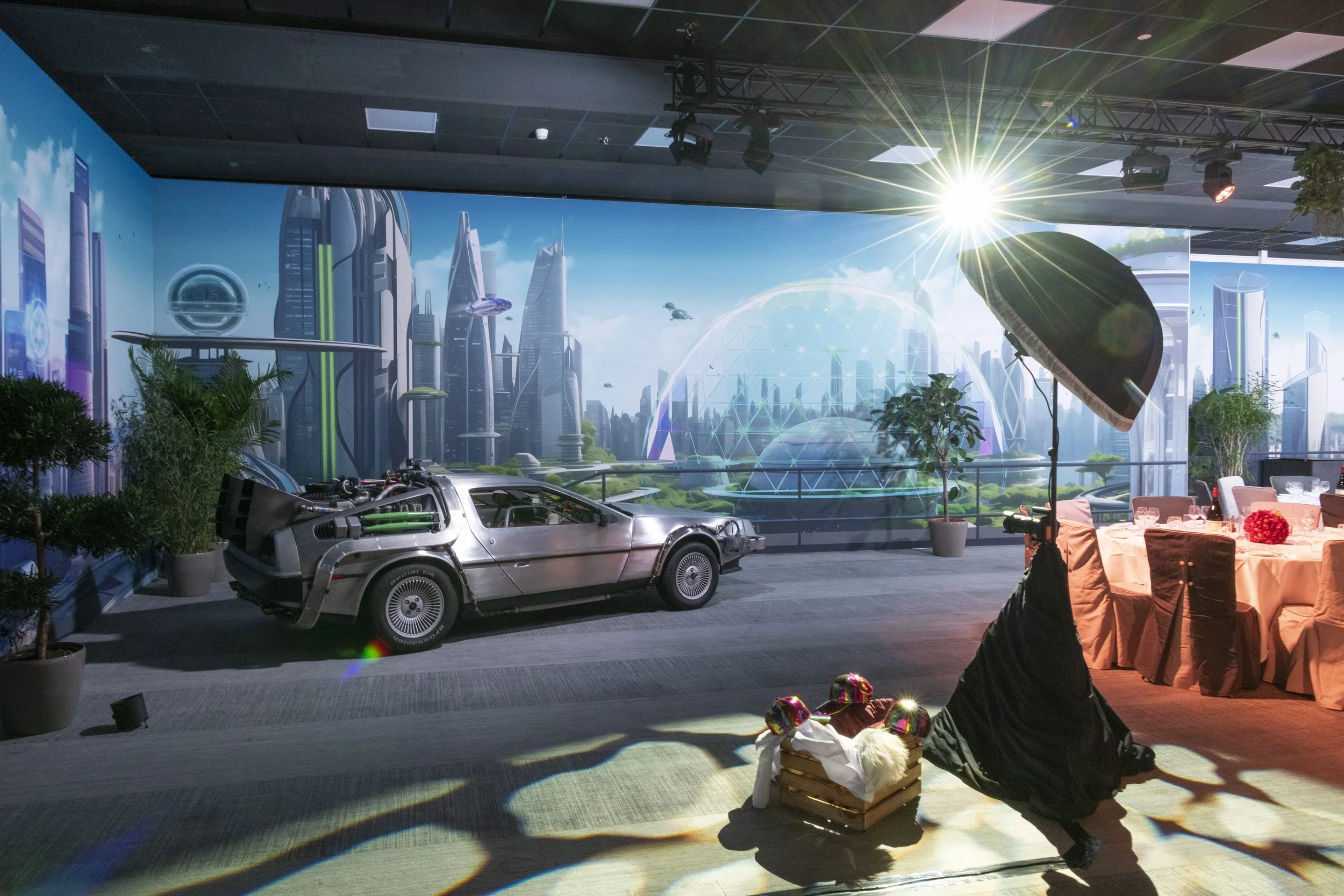 Une scénographie futuriste avec une voiture emblématique de la science-fiction, un fond représentant une ville moderne et un décor de fête avec des tables et des décorations, sous un éclairage simulant un soleil artificiel.