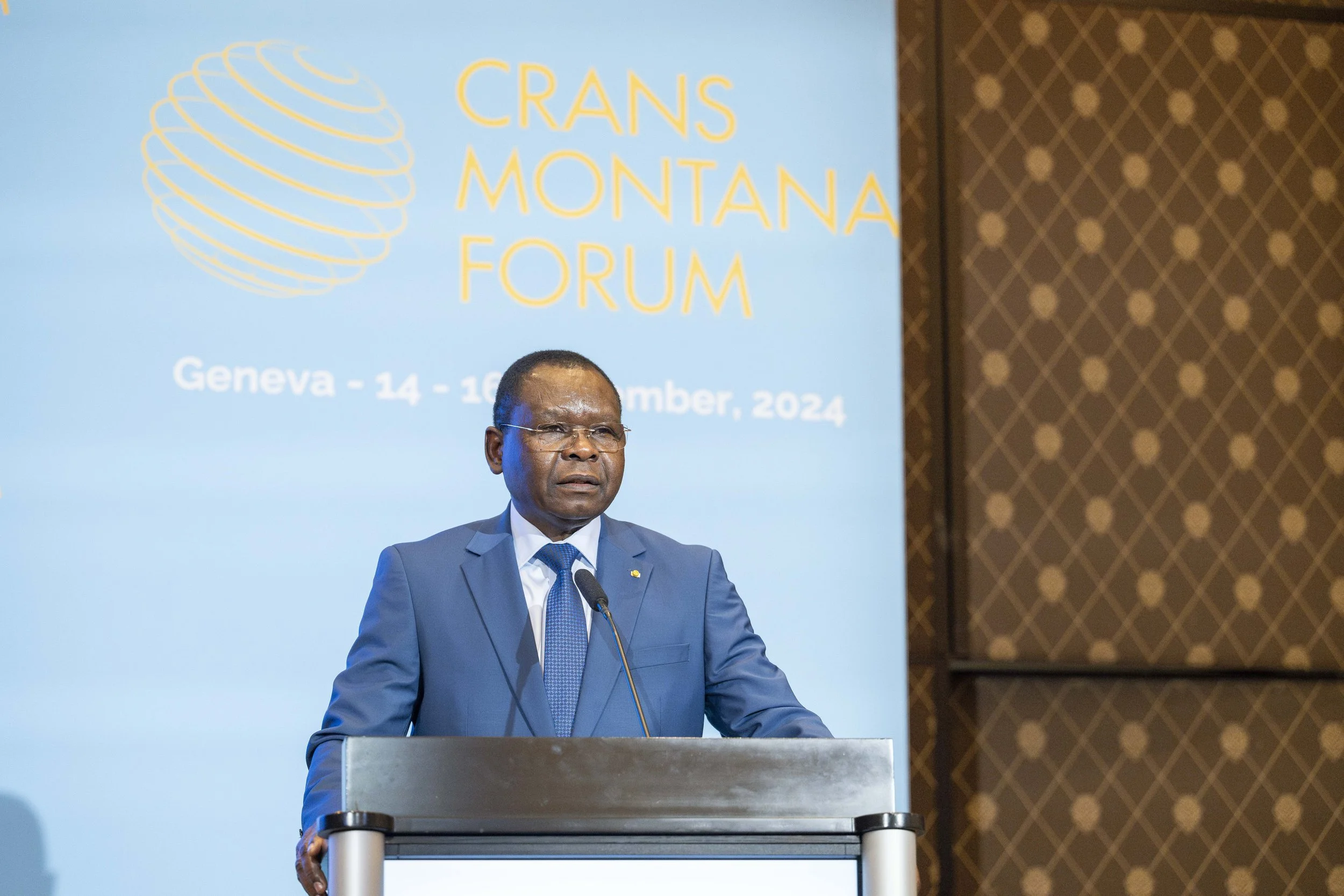 Homme en costume bleu parlant à un podium lors du Crans Montana Forum, avec un panneau bleu en arrière-plan indiquant Genève et les dates du 14 au 16 septembre 2024.