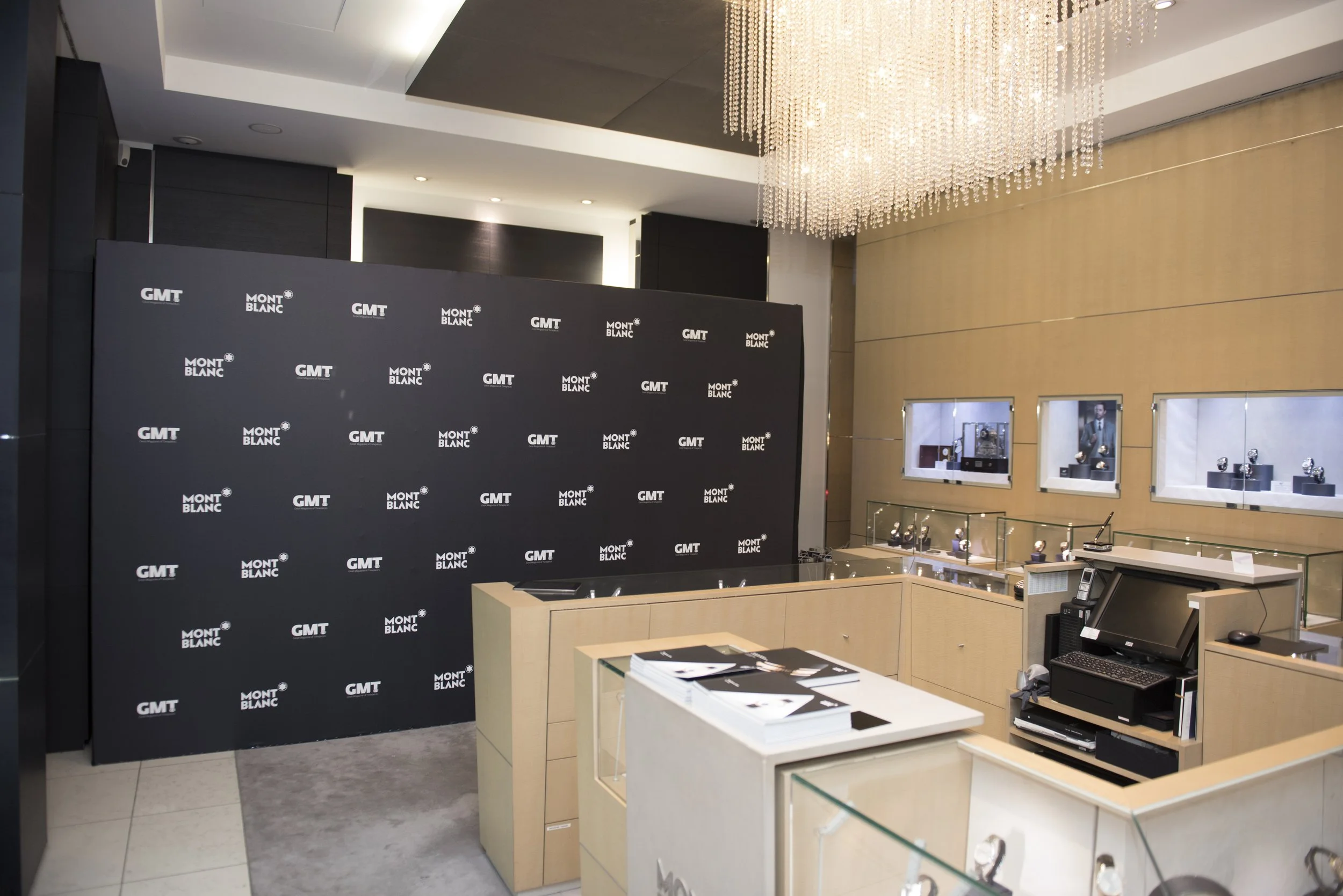 Vitrine avec montres de luxe dans une boutique, fond noir avec logos GMT et Mont Blanc, comptoir en bois clair avec ordinateur et brochures, éclairage sophistiqué, plafond avec chandelier en cristal.