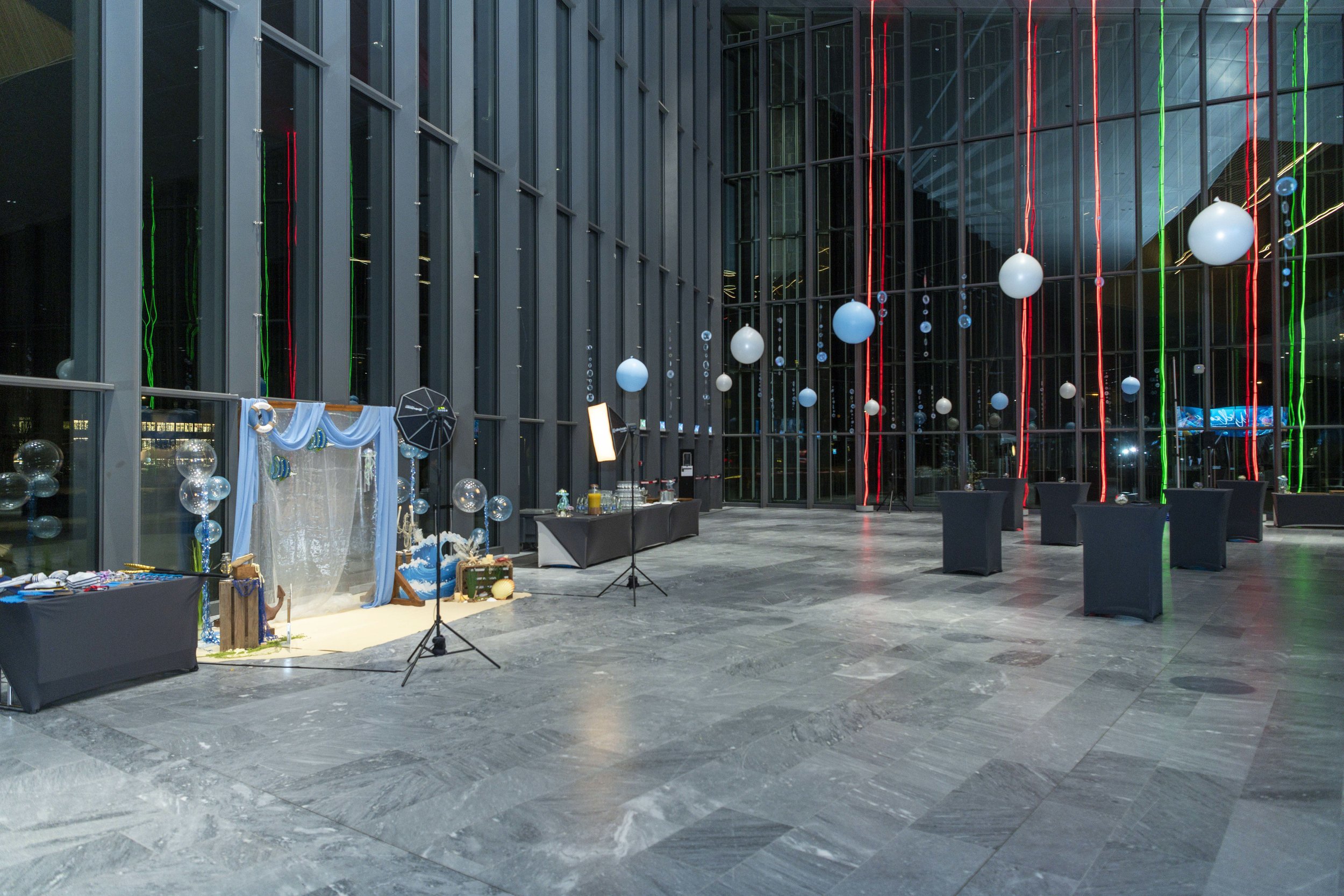 Une salle spacieuse et moderne décorée avec des ballons bleus et blancs suspendus, des bandes lumineuses rouges et vertes, et un coin photobooth avec des accessoires bleus et un éclairage, prête pour une fête ou un événement.