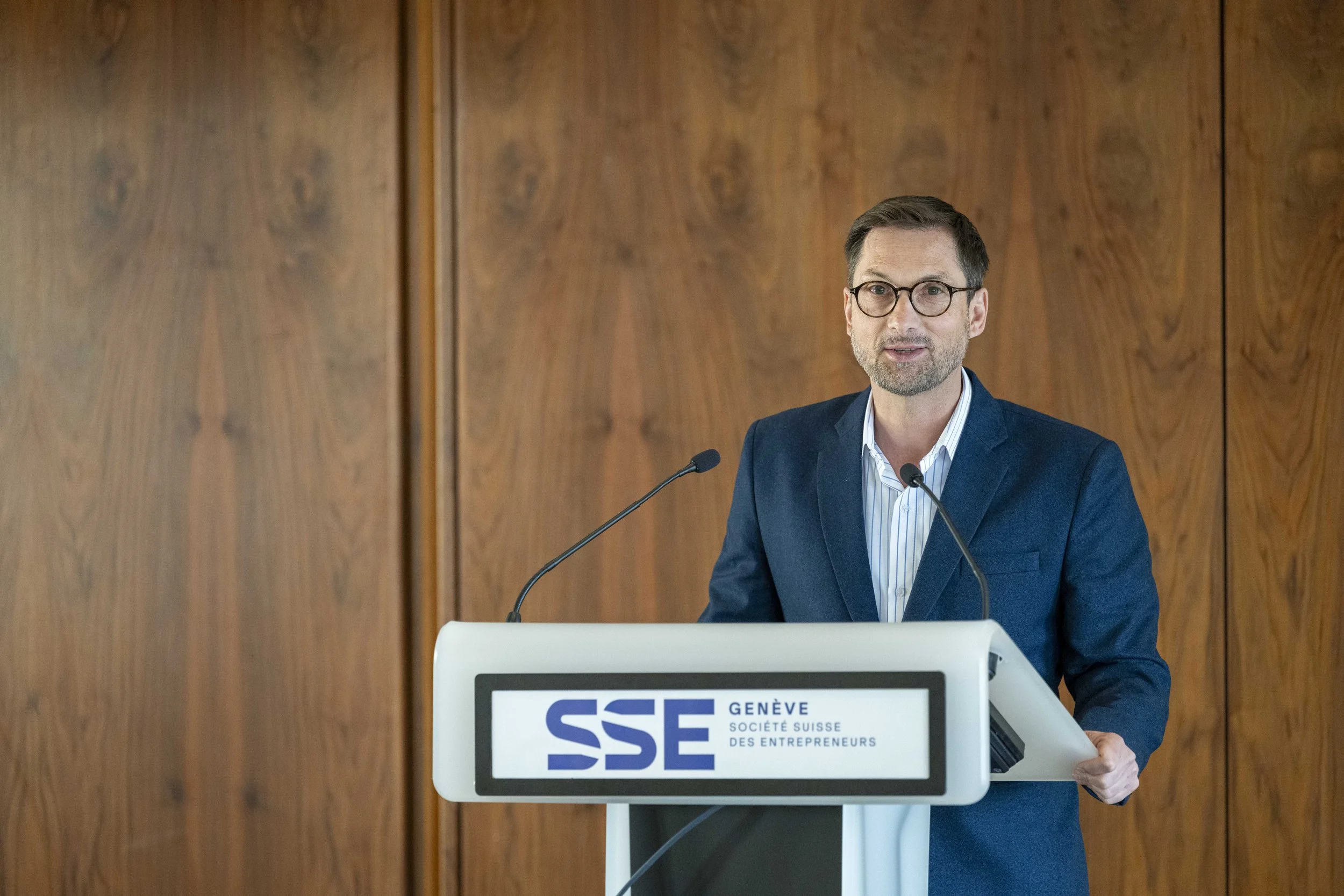 Homme parlant lors d'une conférence derrière un pupitre avec le logo de la SSE Genève et société suisse des entrepreneurs, fond noir, robe bleue, lunettes, fond en bois 