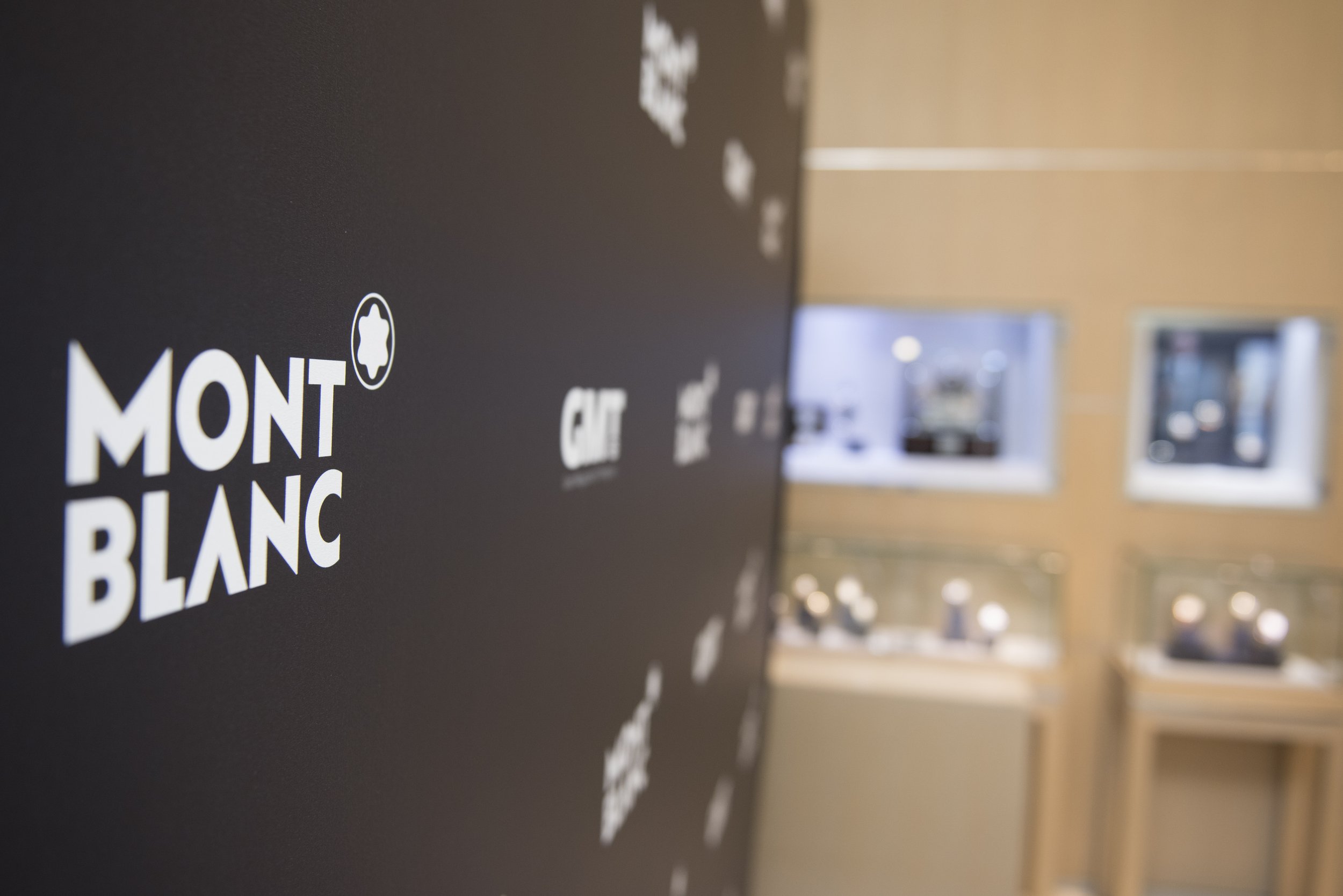 MONTBLANC logo sur un mur noir avec d'autres logos de marques de montres, devant une vitrine avec des montres en arrière-plan.