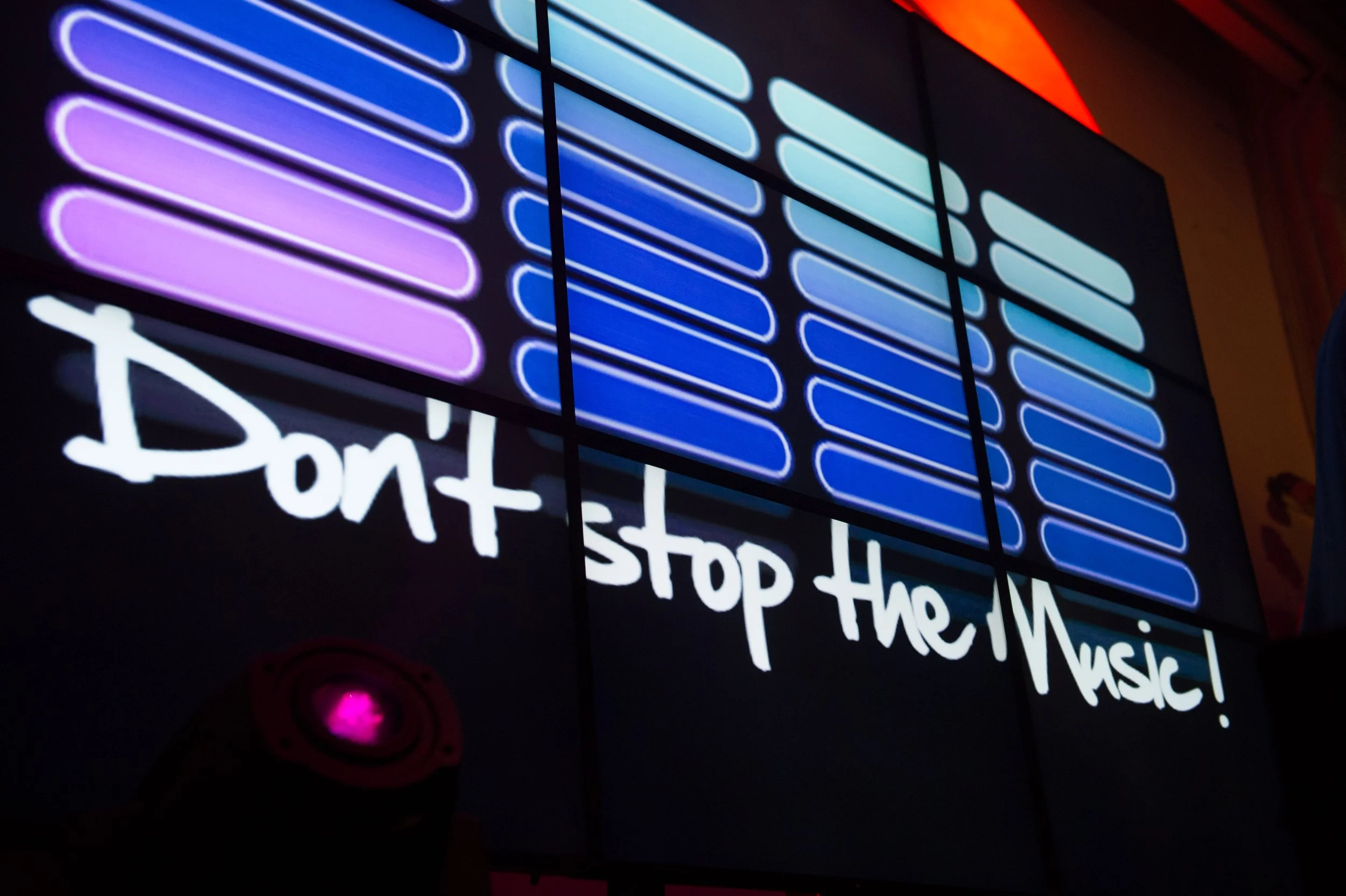 Signal lumineux de style néon avec des barres horizontales dégradées de violet à vert, et des messages en anglais et français concernant le style de musique. La partie visible du message en français dit 'stop the music!'.