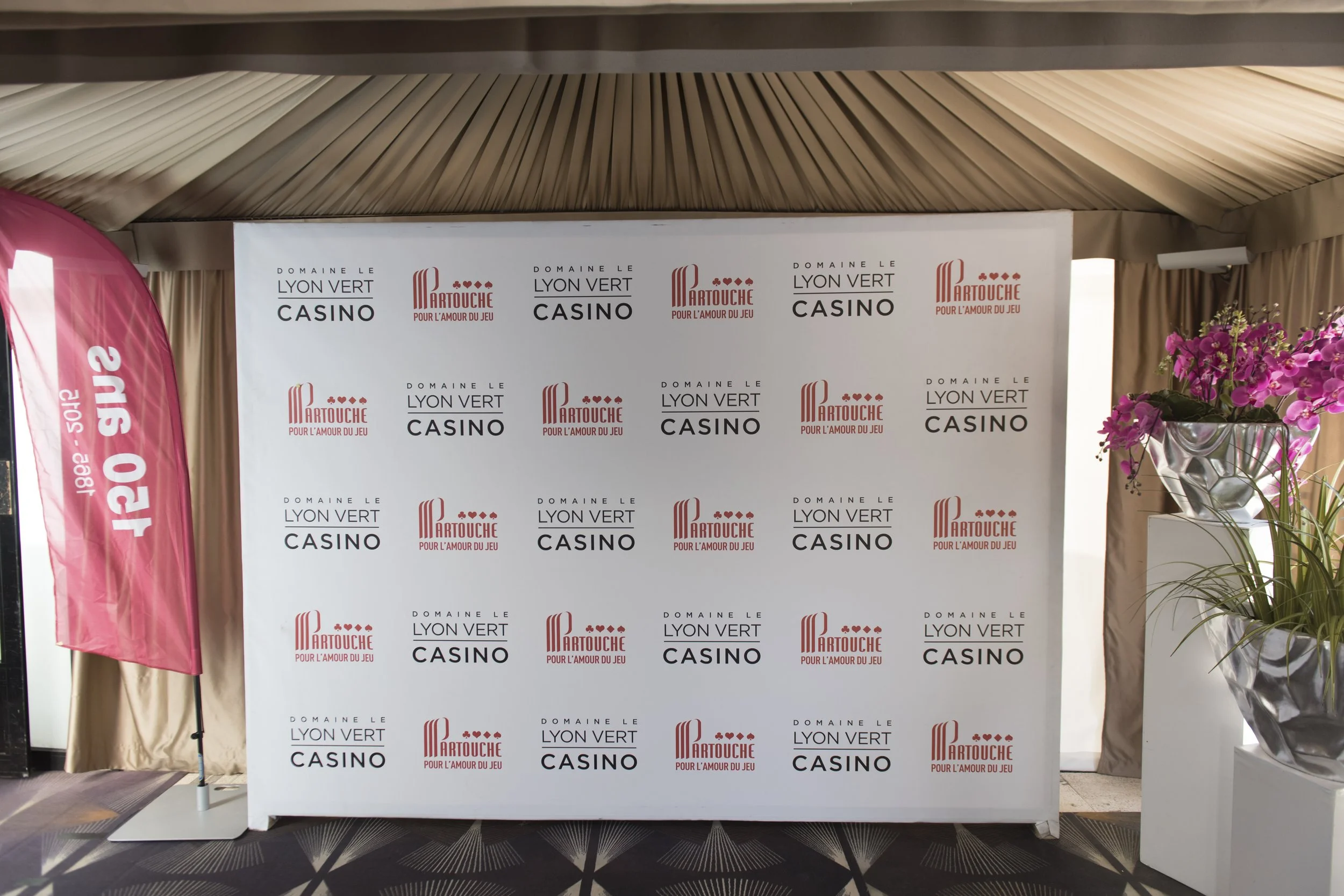 Mur blanc avec logos successifs de la société 'Domaine le Lyon Vert' et 'Partouche' pour un événement de casino, flèche rose à gauche avec texte 'Cjour s'informe' et fleurs violettes dans un vase à droite.