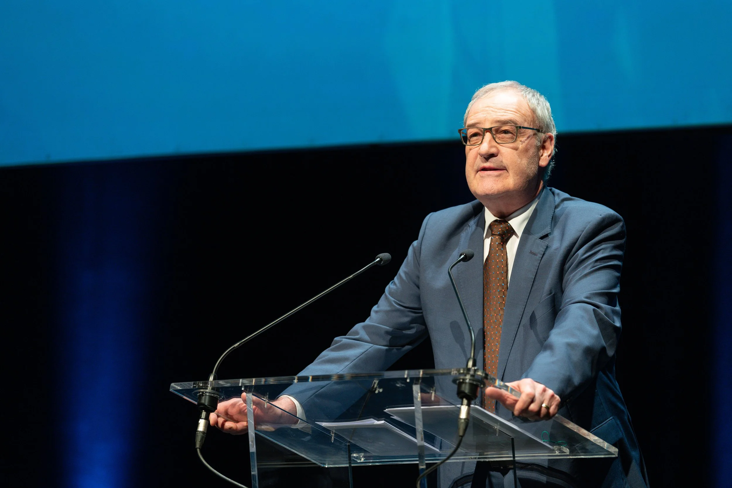 Un homme d'âge moyen portant un costume gris, une chemise blanche et une cravate marron, debout derrière un pupitre avec deux microphones, en train de parler lors d'une conférence ou d'un discours. Il porte des lunettes et a les cheveux gris.