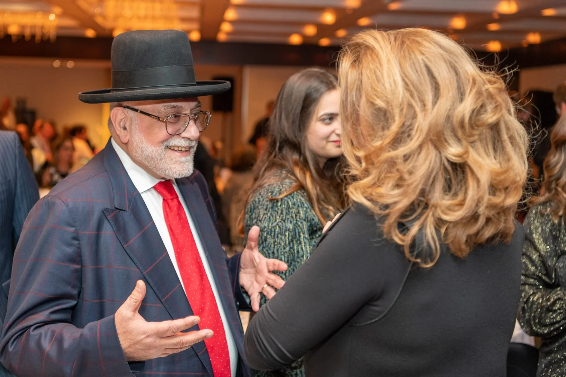Un homme âgé avec une barbe grise, portant un chapeau noir, des lunettes, une veste à carreaux, une chemise blanche et une cravate rouge, sourit en discutant avec une femme aux cheveux roux et longs, portant une robe noire, dans une salle de réceptio