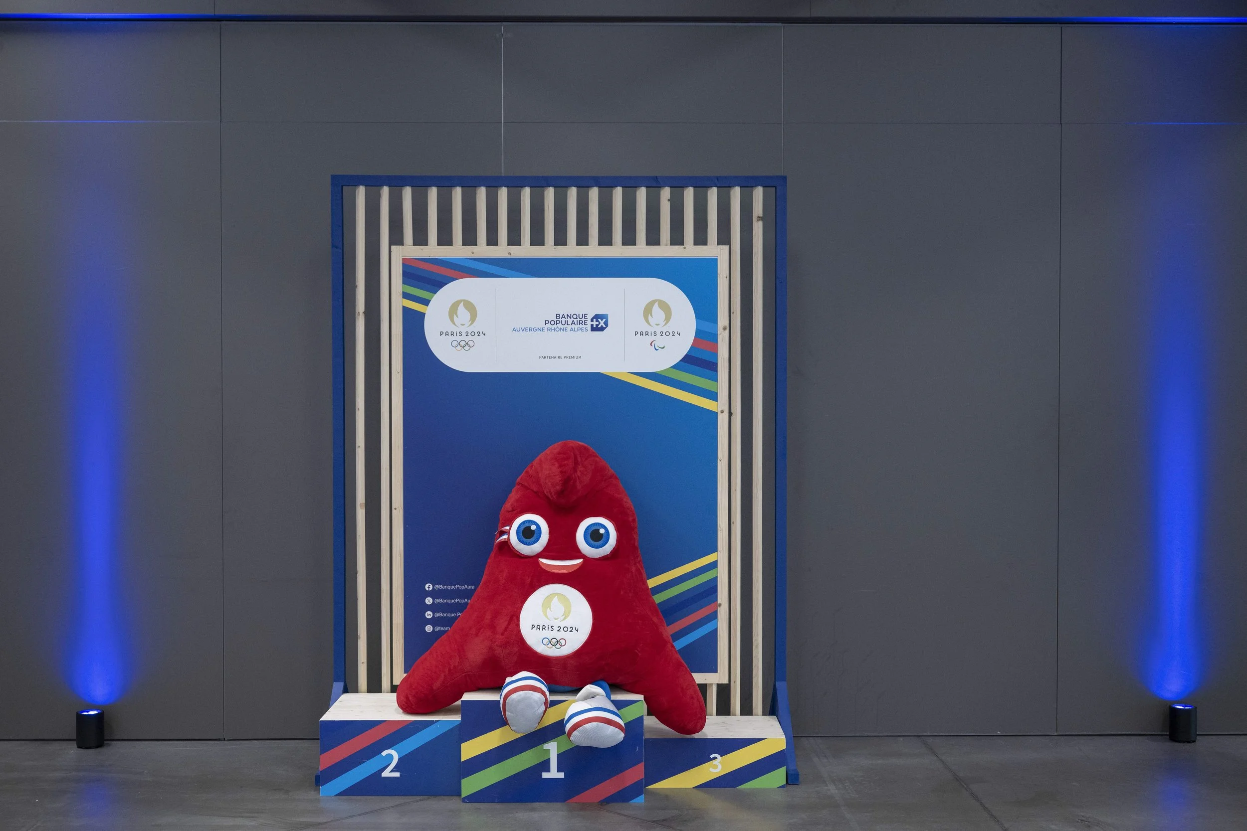 Une mascotte en forme de point rouge avec de grands yeux, un sourire, et portant un badge avec le logo des Jeux Olympiques de Paris 2024, est assise devant un podium avec les places 1, 2 et 3, dans une salle avec un mur gris et des éclairages bleus.