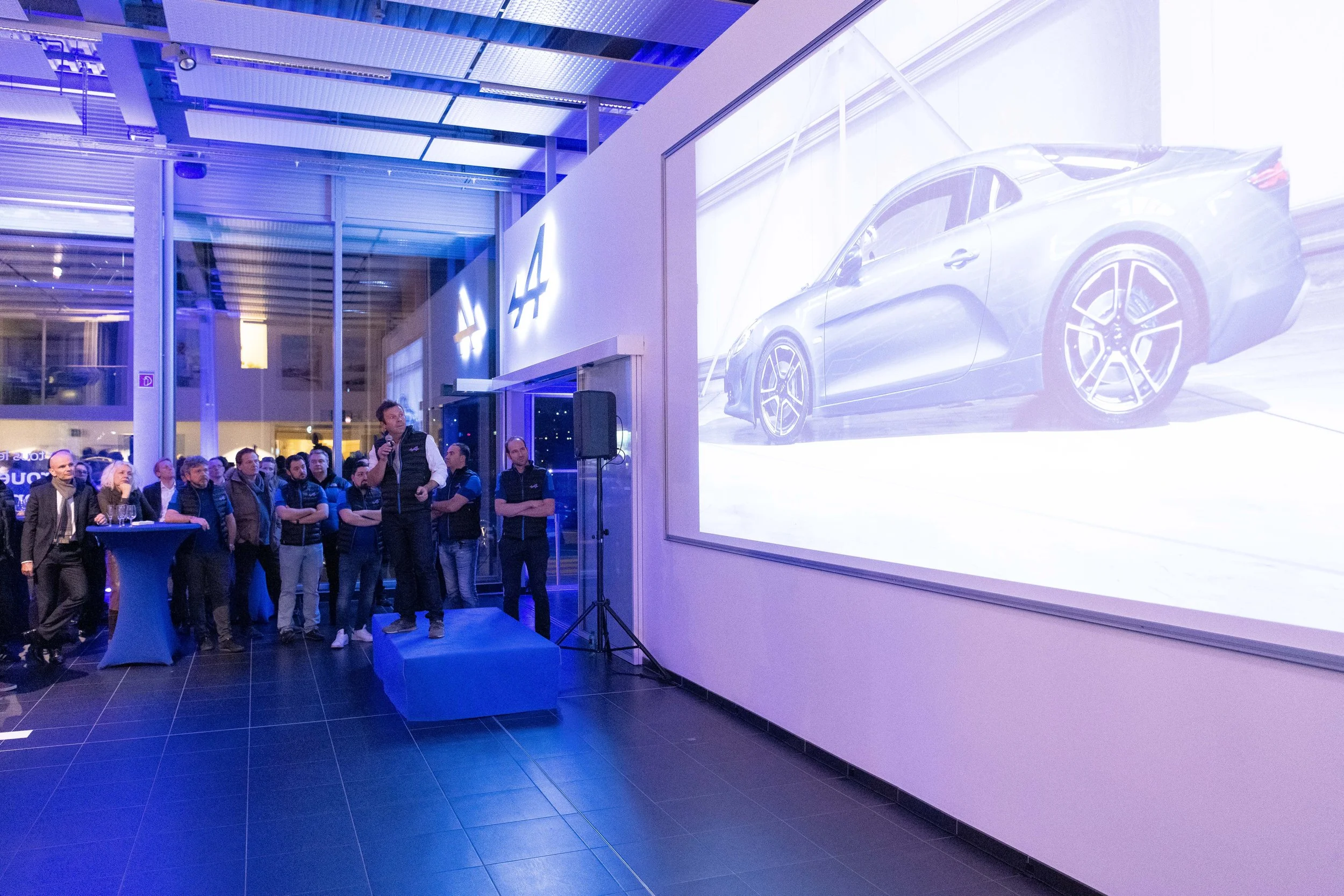 Une présentation en intérieur avec un groupe de personnes écoutant un homme parler, devant une grande image d'une voiture sur un écran.