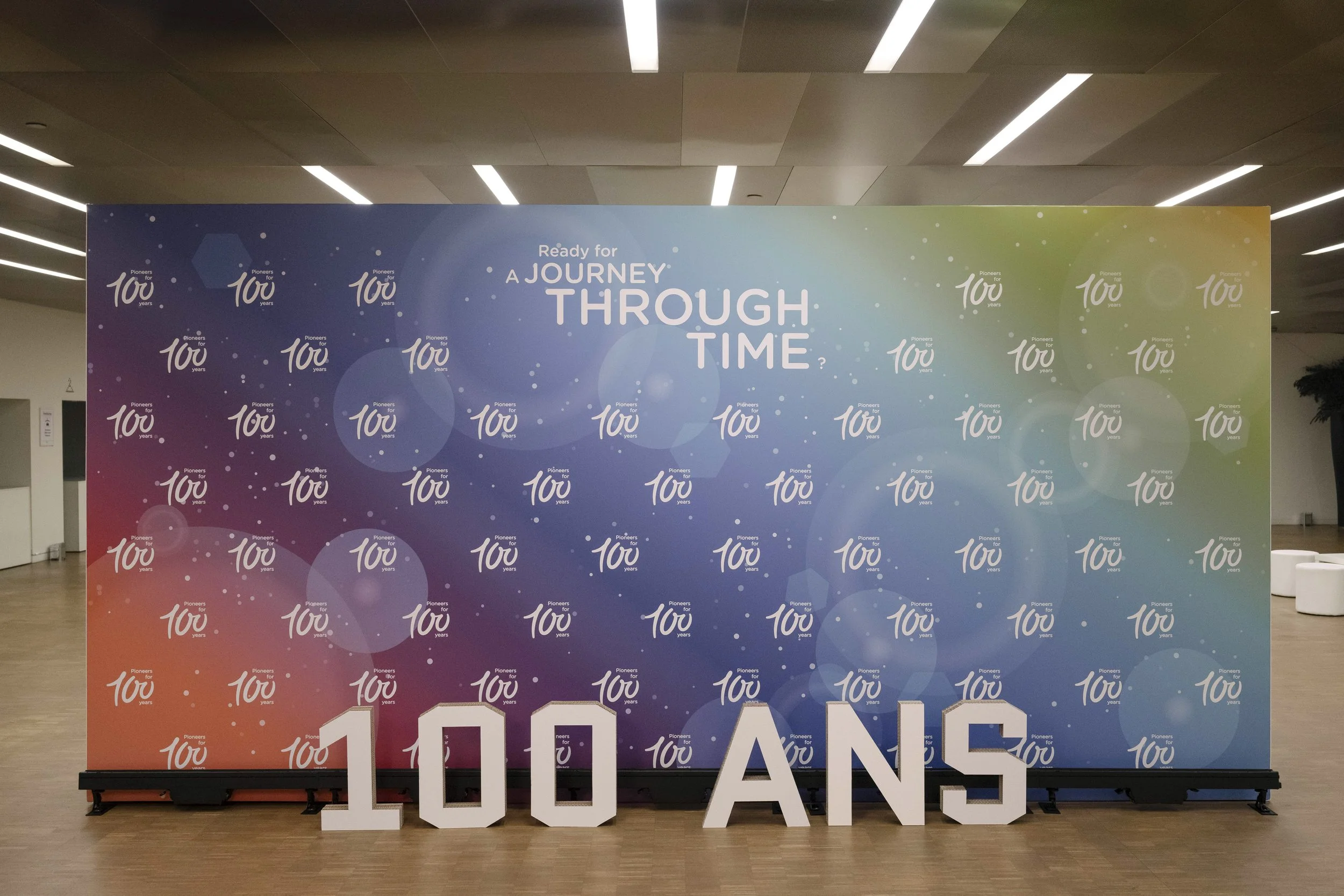 Grand mur avec un fond coloré dégradé de bleu, vert, jaune et orange, avec le texte 'Ready for a journey through time'. En bas, de grands chiffres blancs formant '100 ANS'.