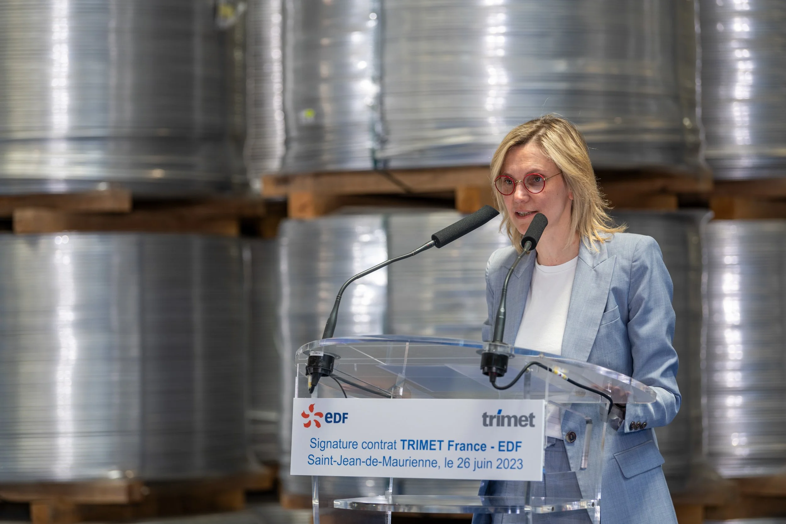 Une femme parle lors d'une conférence de presse devant un podium avec un panneau indiquant « Signature contrat TRIMET France - EDF » le 26 juin 2023, dans une salle avec des bobines métalliques en arrière-plan.