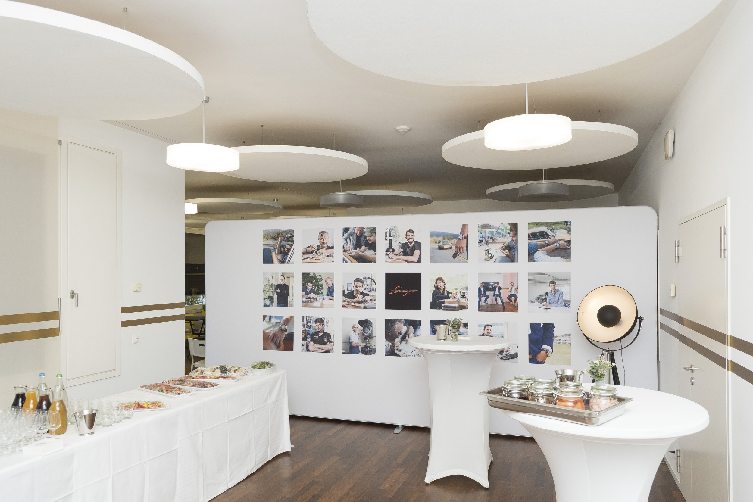 Une salle d'exposition ou de réception avec un panneau d'affichage composé de plusieurs photos de personnes, un buffet avec des boissons et des plats, ainsi que des tables avec des décorations. 