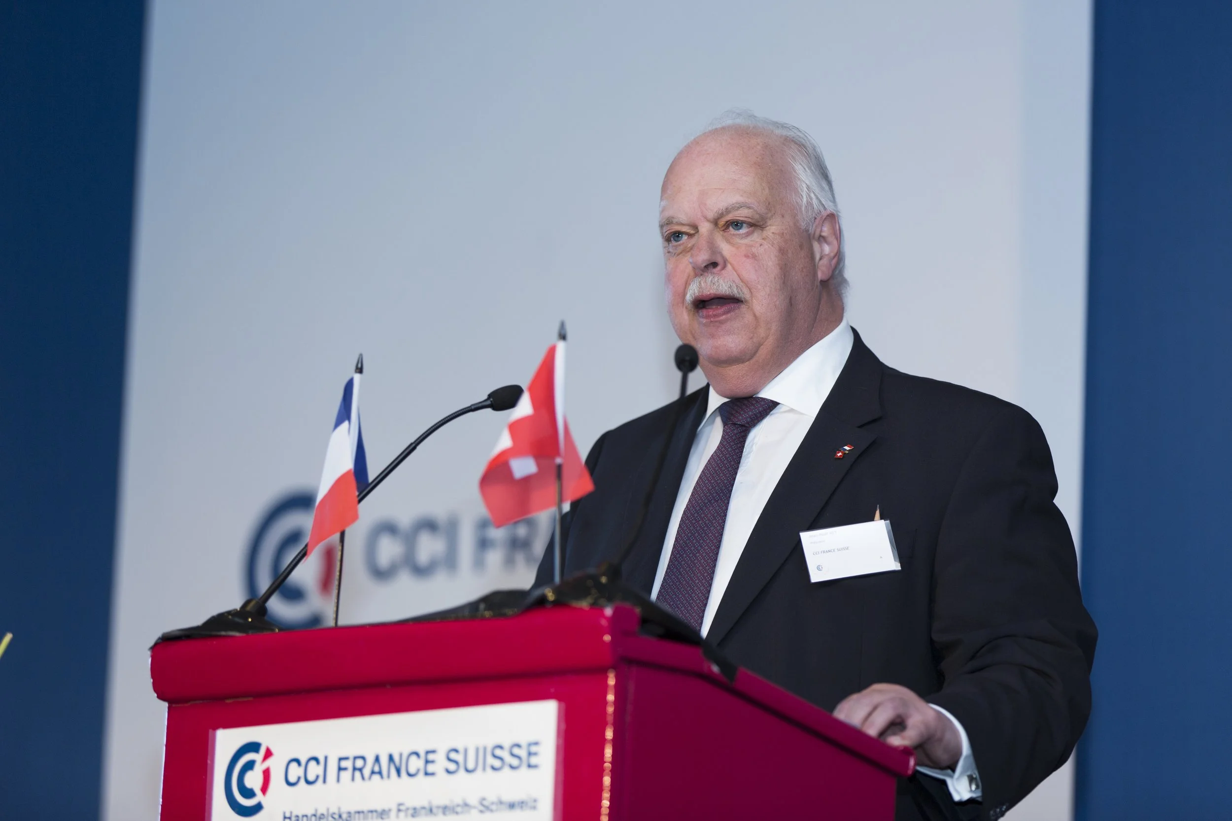 Homme en costume noir et cravate, parlant lors d'une conférence, avec deux micros, devant un fond blanc avec le logo CCI France Suisse, à côté de deux drapeaux, un suisse et un français.