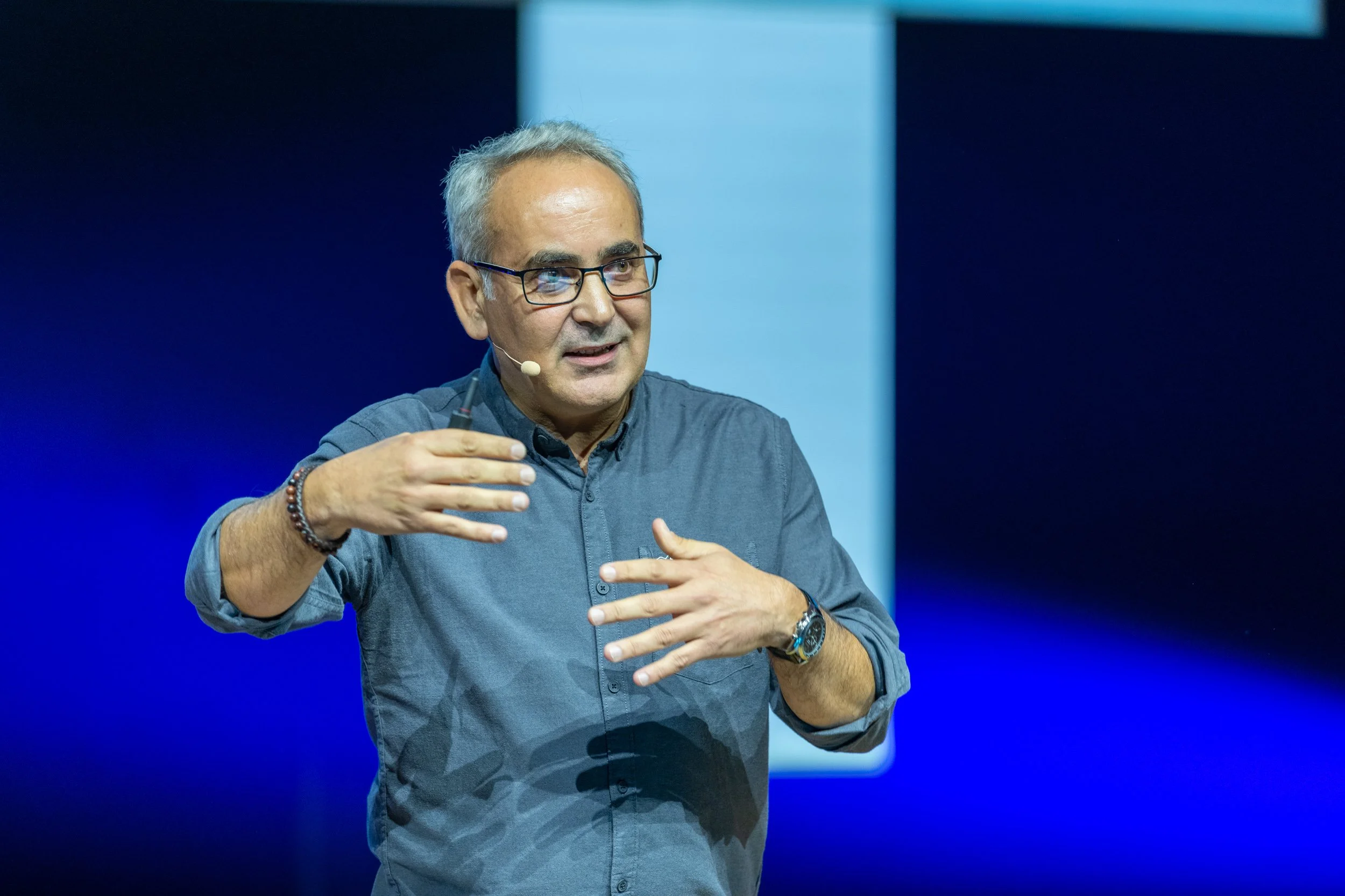 Un homme parlant lors d'une conférence, portant une chemise grisâtre, avec un micro-cravate et des lunettes, gesticulant avec ses mains.
