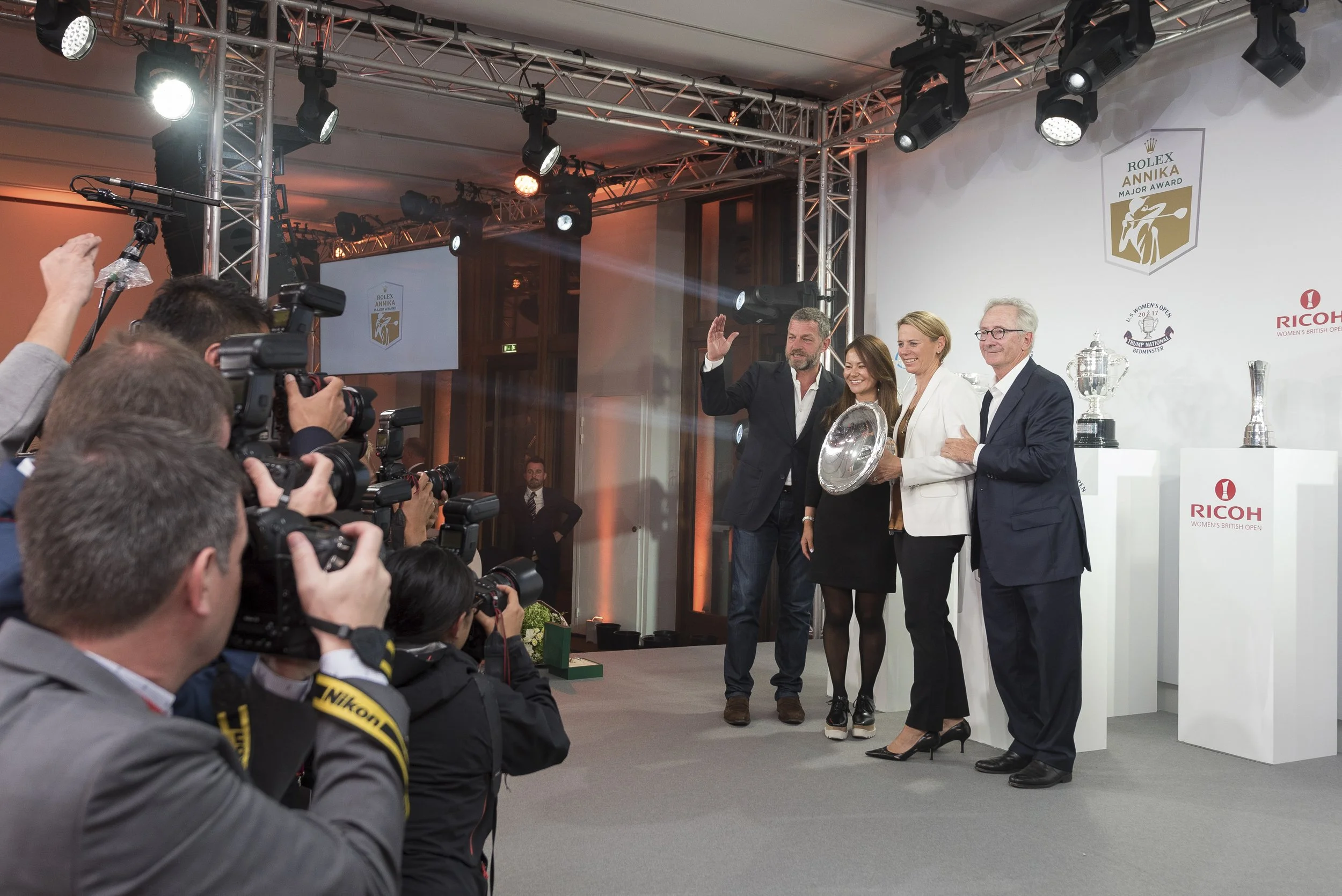 Cérémonie de remise de prix lors d'un événement sportif, avec plusieurs personnes sur scène tenant un trophée ou un plat en argent, entourées de photographes. La scène est équipée d'un système d'éclairage professionnel et affiche des logos de la comp