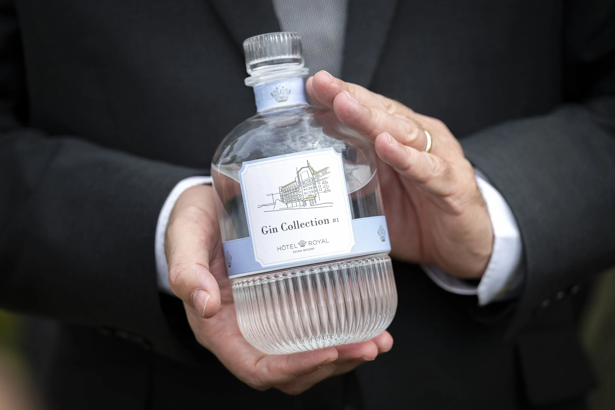 Une personne en costume tient une bouteille d'alcool appelée 'Gin Collection #1' du Hôtel Royal, avec un dessin d'hôtel sur l'étiquette.