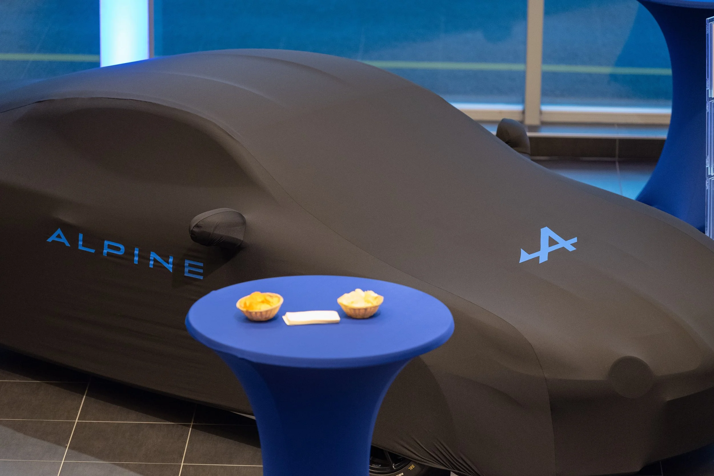 Une voiture de course compacte, grise, avec le mot 'ALPINE' en bleu et un logo blanc, exposée dans un espace intérieur, avec une petite table bleue devant elle contenant deux assiettes de nourriture et des serviettes.