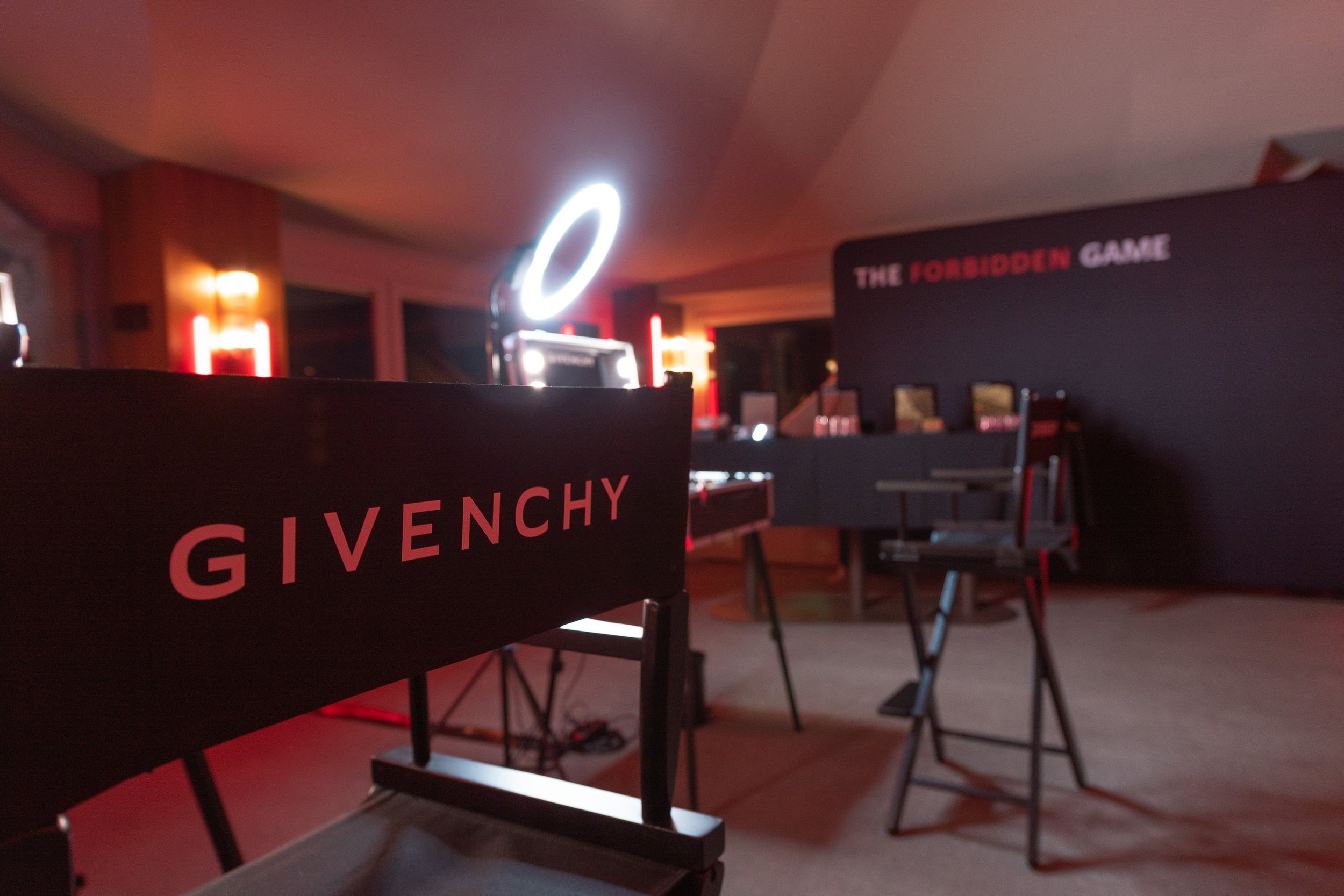 Salle sombre avec une table et des chaises, décorée pour un jeu ou un événement, avec une enseigne 'The Forbidden Game' et une bannière 'Givenchy' en avant-plan.