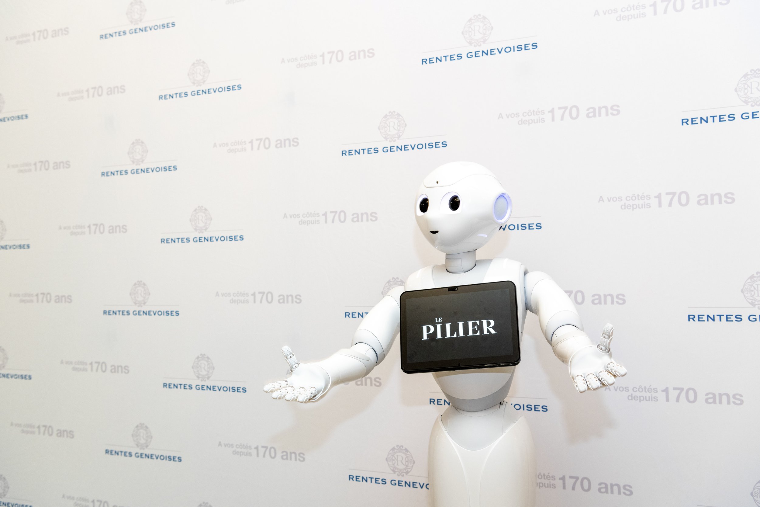 Un robot humanoïde blanc avec un écran sur la poitrine affichant 'LE PILIER' se trouve devant un mur blanc avec des logos et du texte en français, avec des bras ouverts.
