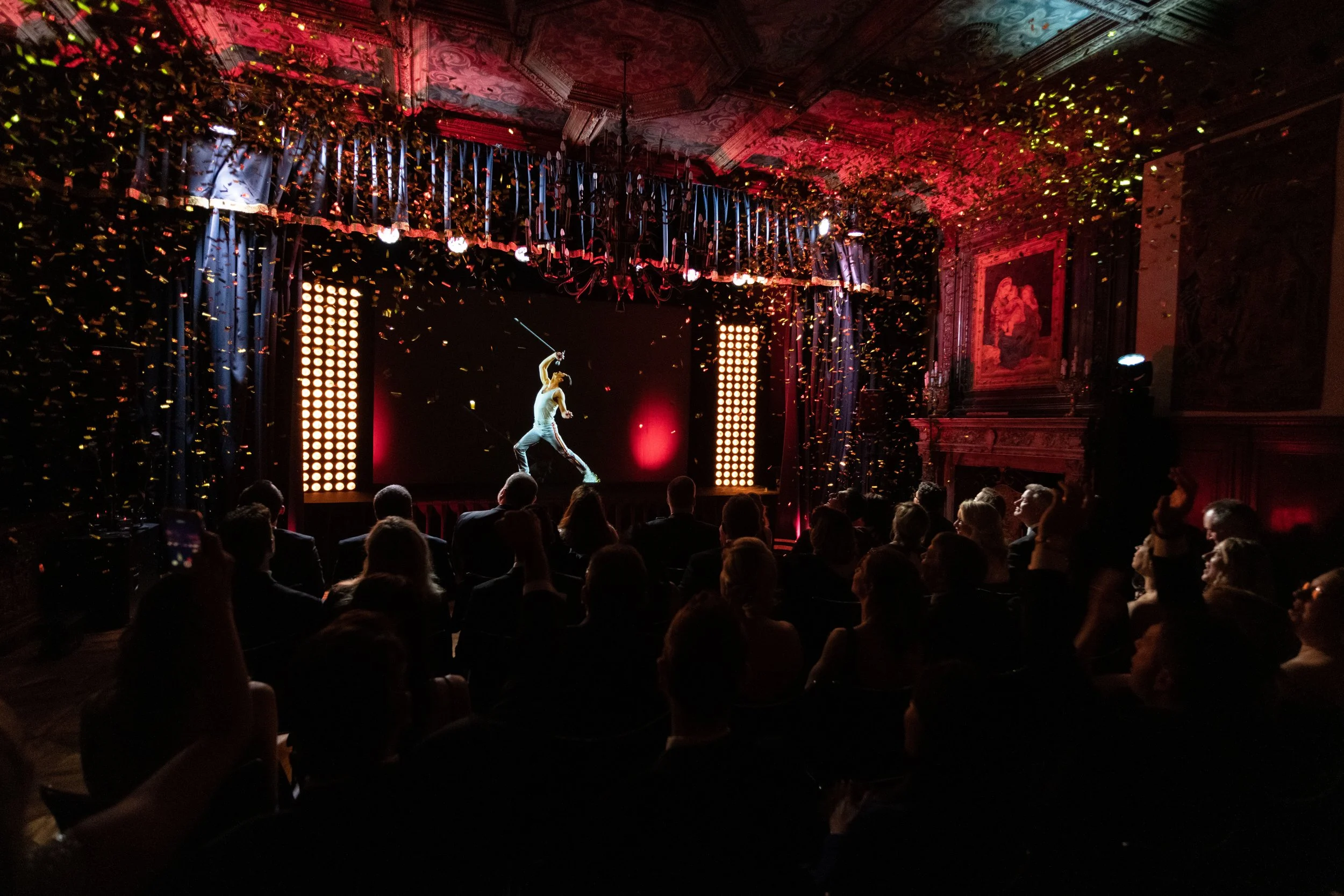 Une scène de spectacle avec deux artistes en costume de cirque sur un écran au fond, entourée de confettis dorés en suspension, avec un public en silhouette assis dans une salle ornée de décorations et de rideaux bleus.