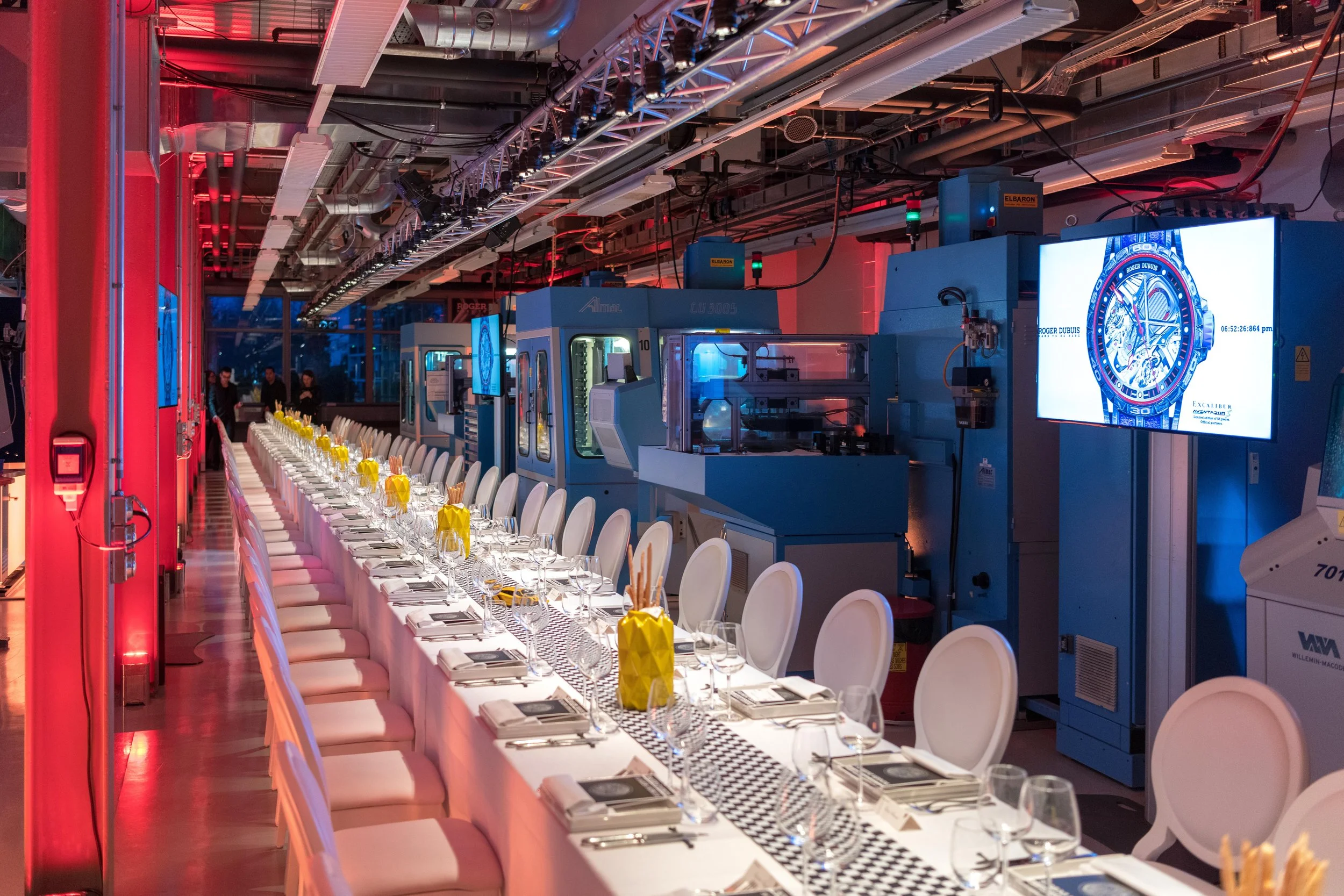 Une longue table de dîner élégante avec nappes blanches, assiettes, verres à vin, et petites décorations jaunes, dans une salle moderne avec des machines industrielles et un grand écran affichant une montre.