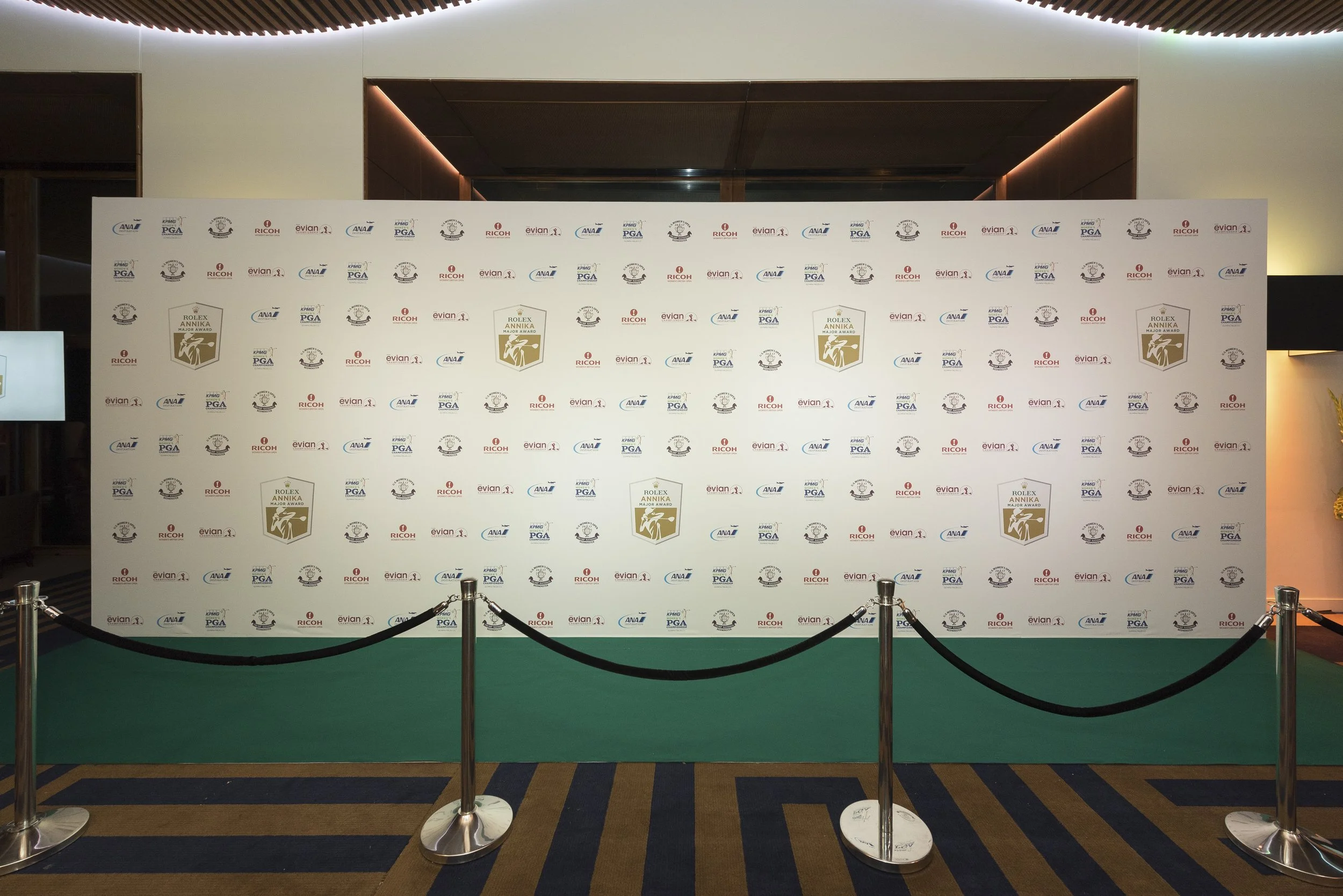 Un tapis vert et une barrière noire entourent un panneau blanc avec plusieurs logos, dont ceux de la PGA, Ricoh, Evian, et Rolex, suggérant une zone réservée pour un événement sportif ou de remise de prix.