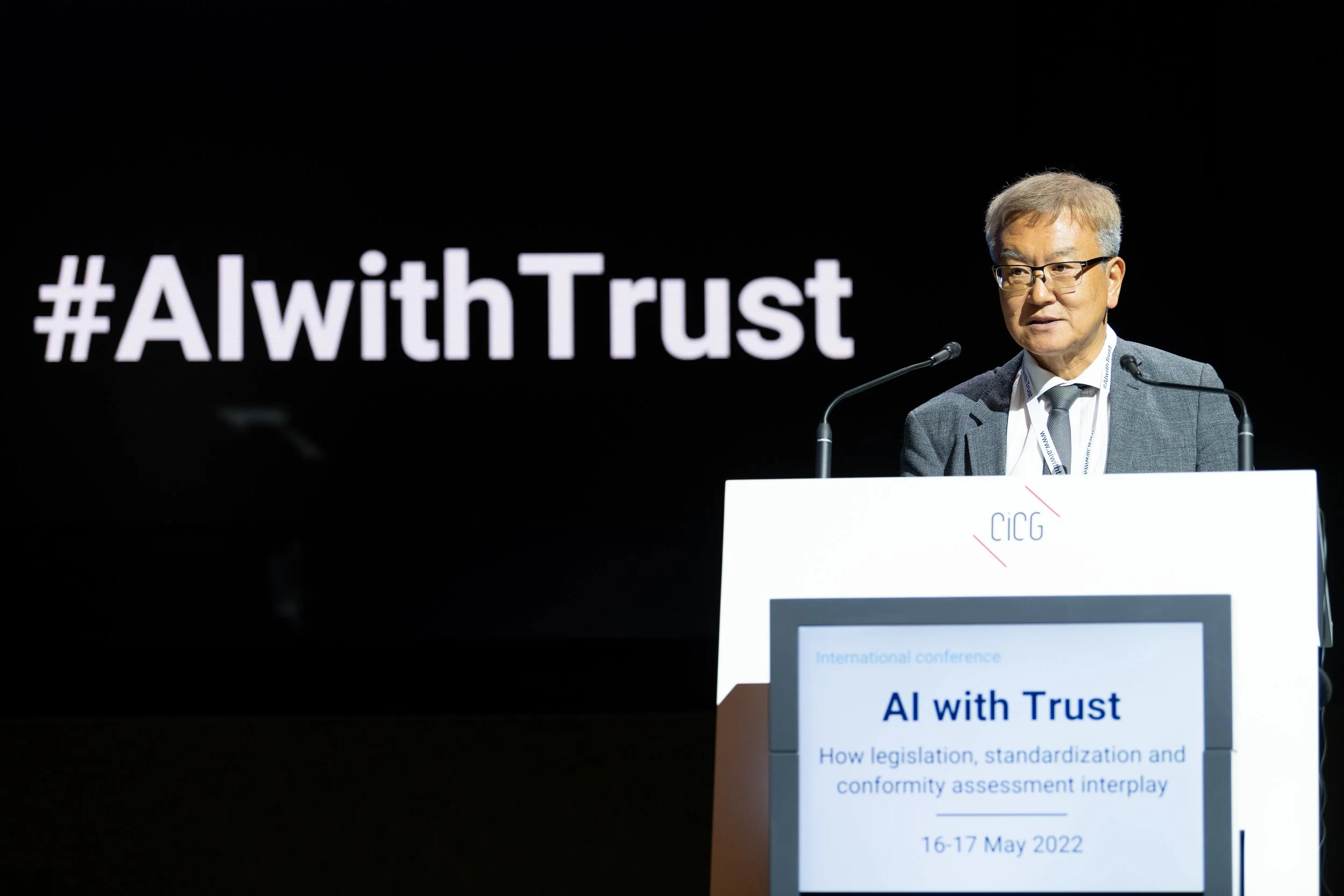 Un homme en costume donne une conférence devant un écran noir avec le hashtag #AIwithTrust écrit en blanc. La présentation à l'écran indique le titre "AI with Trust" et mentionne la date du 16-17 mai 2022.