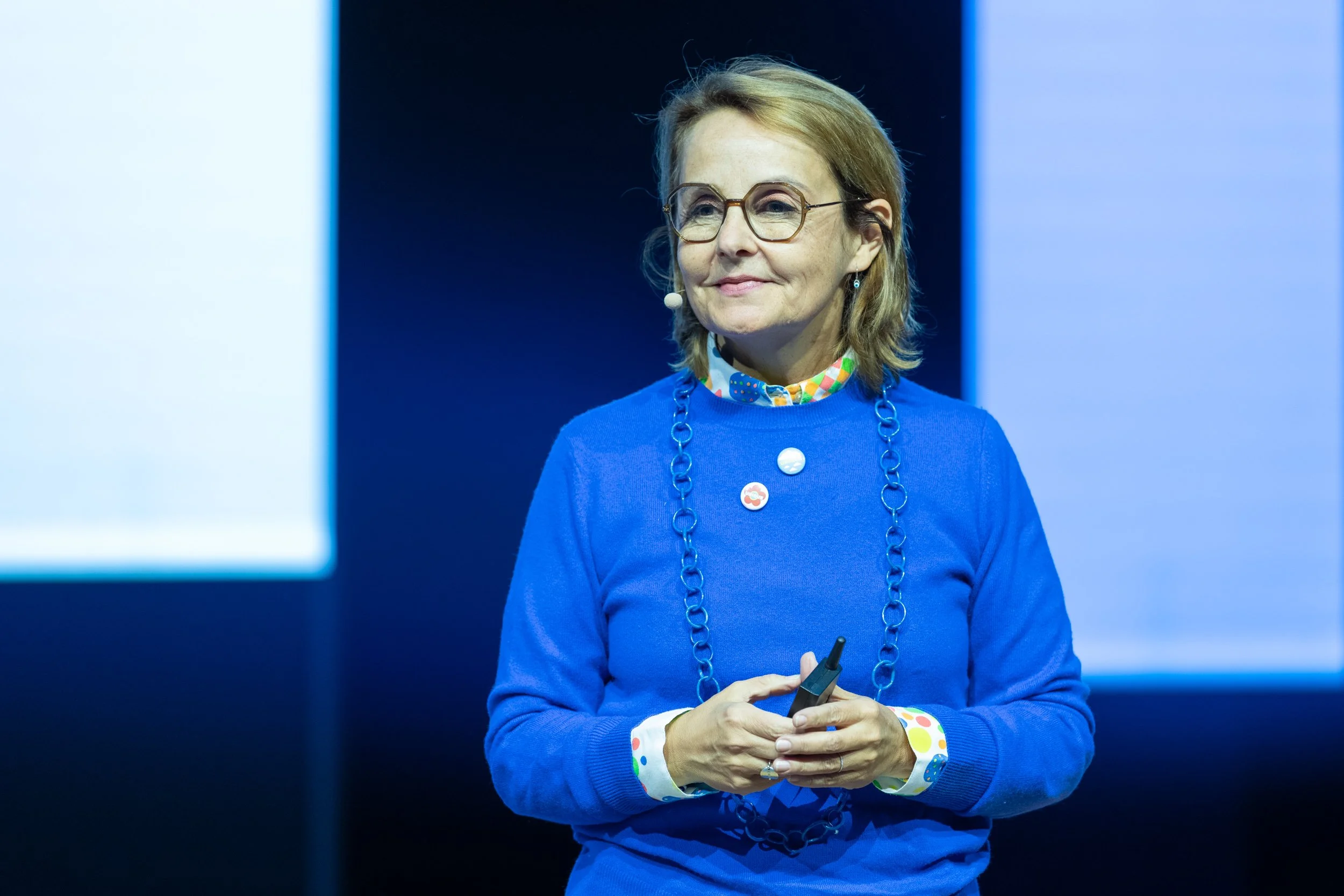 Une femme porte un pull bleu avec un collier et bouton colorés, des boutons à motifs sur la chemise, des lunettes, et tient un micro, en train de parler ou de présenter lors d'un événement. En arrière-plan, un écran lumineux.