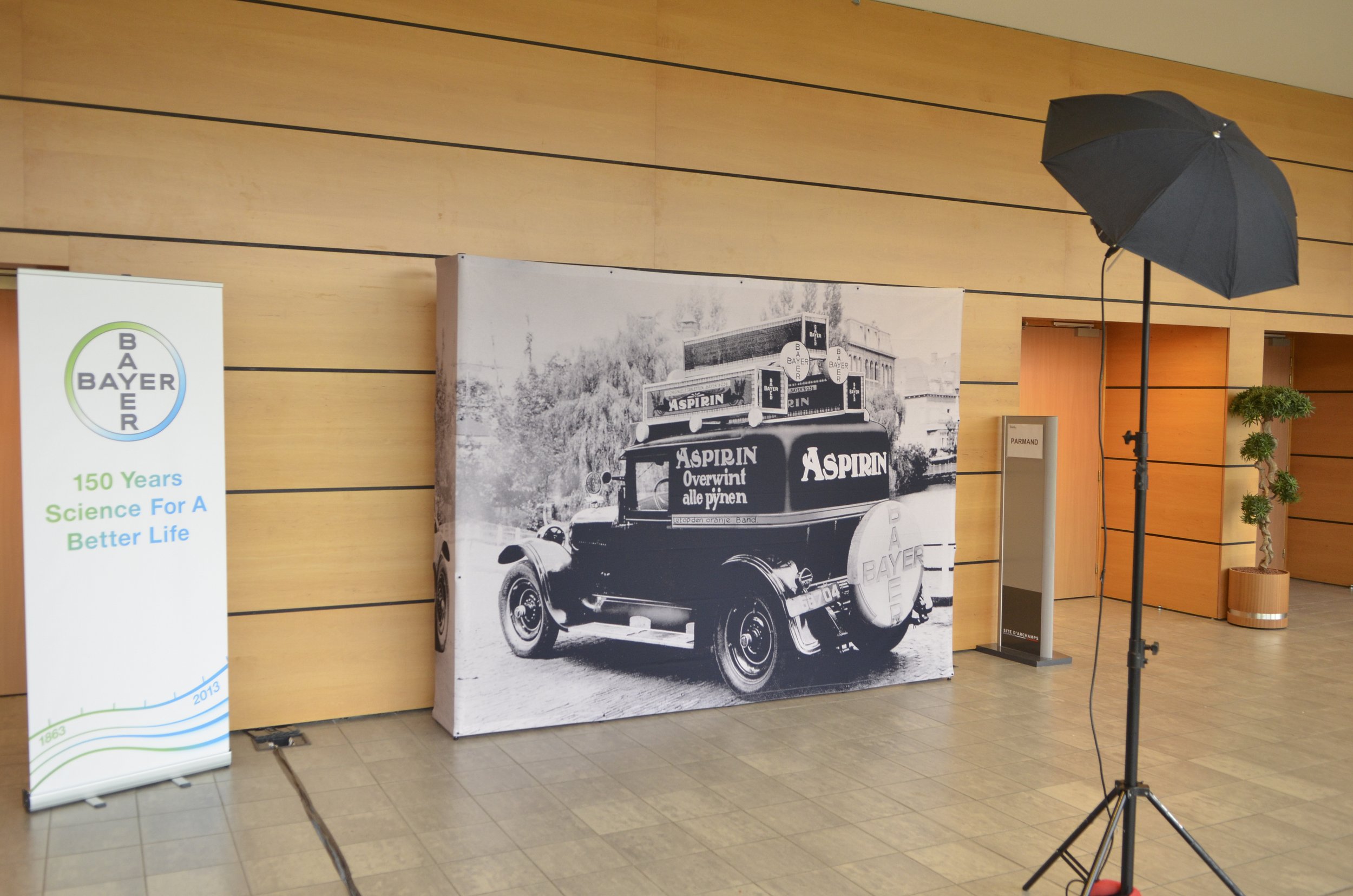 Une étagère de feu qui présente un panneau d'affichage avec un vieux véhicule affiché dessus, décorée avec des produits Bayer, et un drapeau promotionnel à gauche. À droite, un parapluie d'éclairage et une petite plante verte en pot dans une pièce av