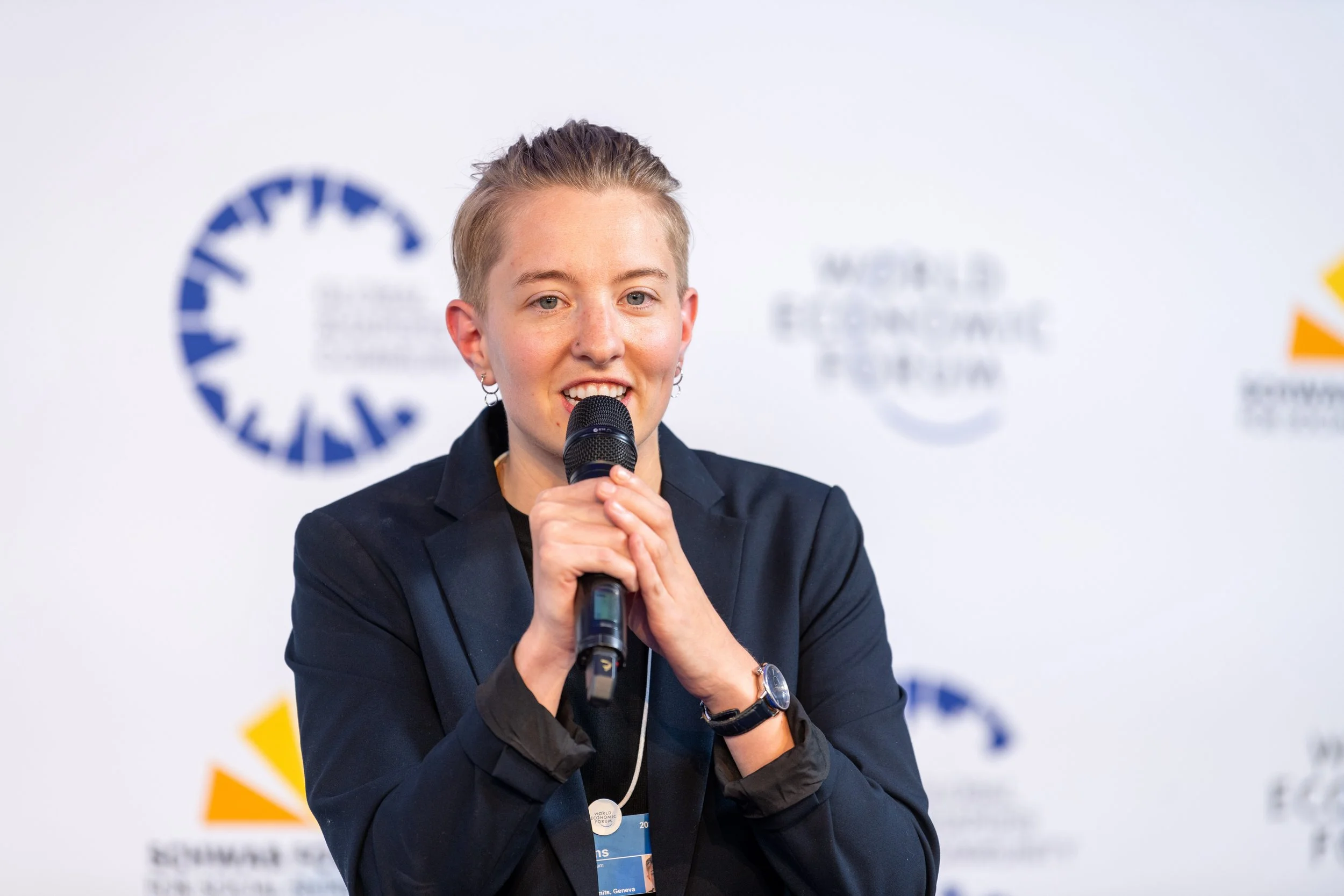 Femme tenant un microphone et parlant lors d'une conférence, avec un fond blanc et des logos d'événements internationaux.