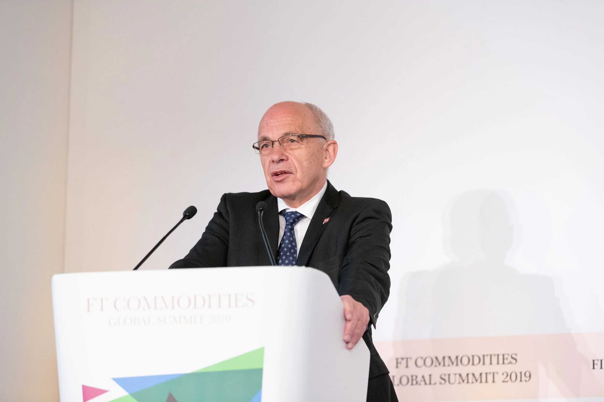 Un homme en costume noir, portant une paire de lunettes, parle lors d'une conférence au sommet mondial des matières premières 2019, derrière un pupitre blanc avec le logo "FT COMMODITIES".