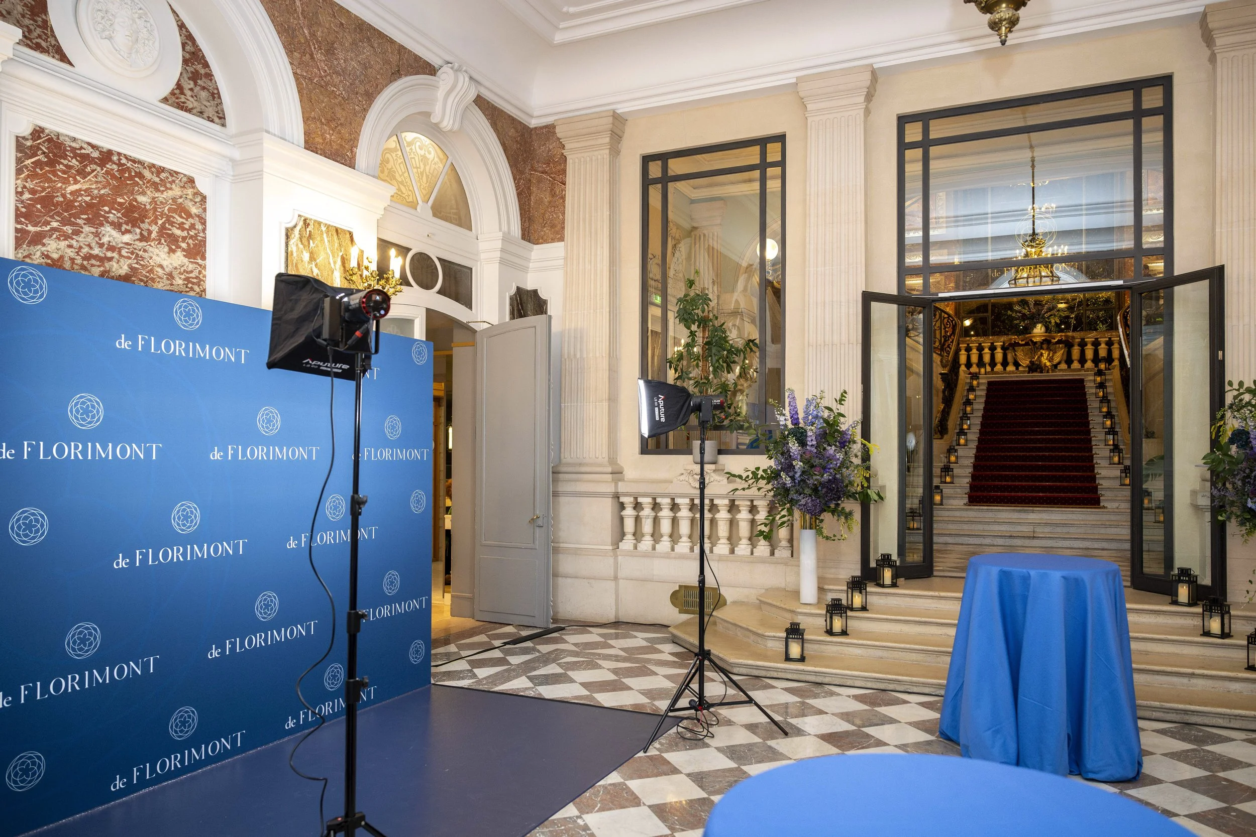 Intérieur élégant avec escaliers en marbre, grande porte en verre, fleurs dans des vases, tables avec nappes bleues, et équipement photo pour une cérémonie ou un événement.