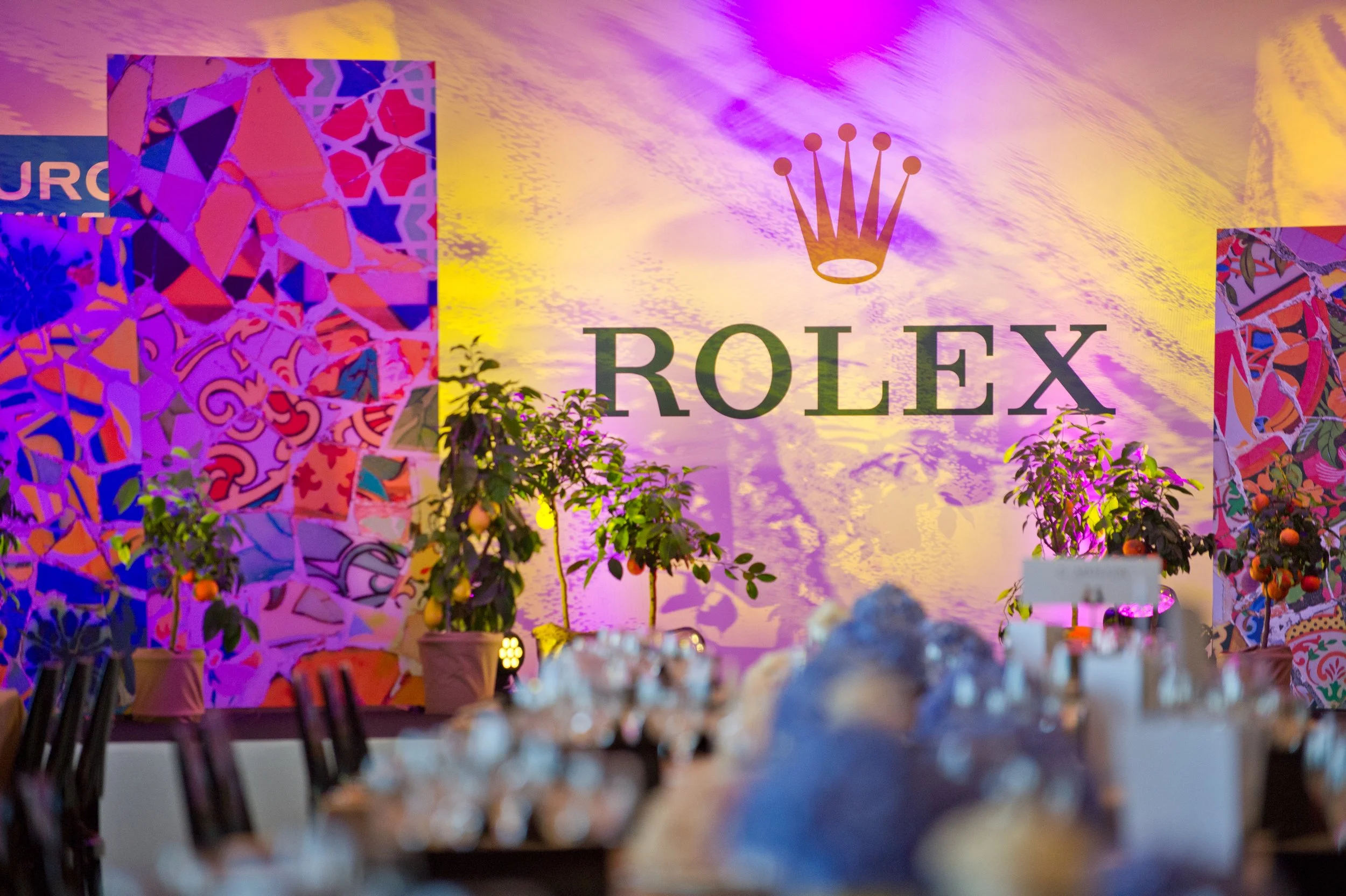 Une scène d'un événement Rolex avec un grand panneau arborant le logo Rolex et un fond coloré en violet et jaune, entourée de décorations colorées et de petits arbres, avec des tables à l'avant et des objets en verre sur la table.