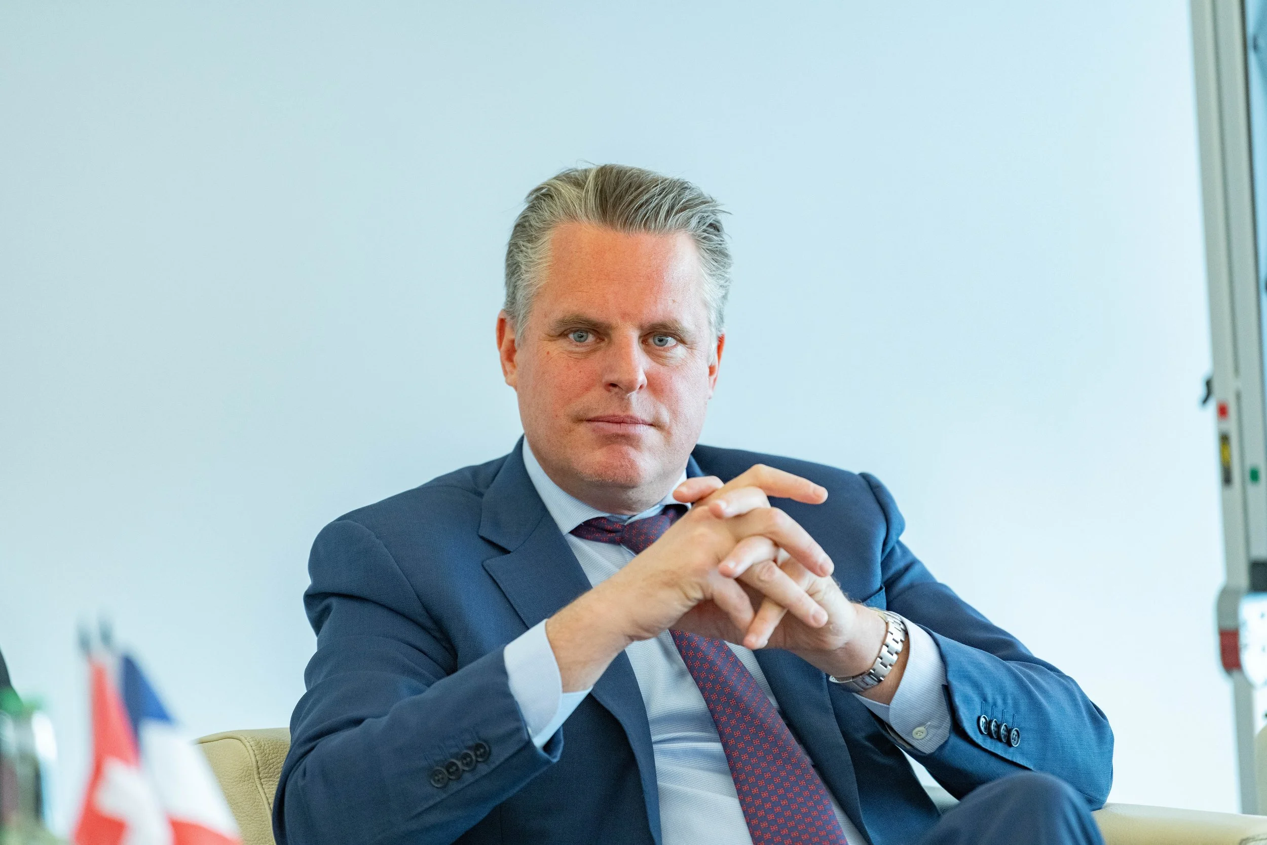 Homme en costume bleu, assis avec mains jointes, dans un bureau.