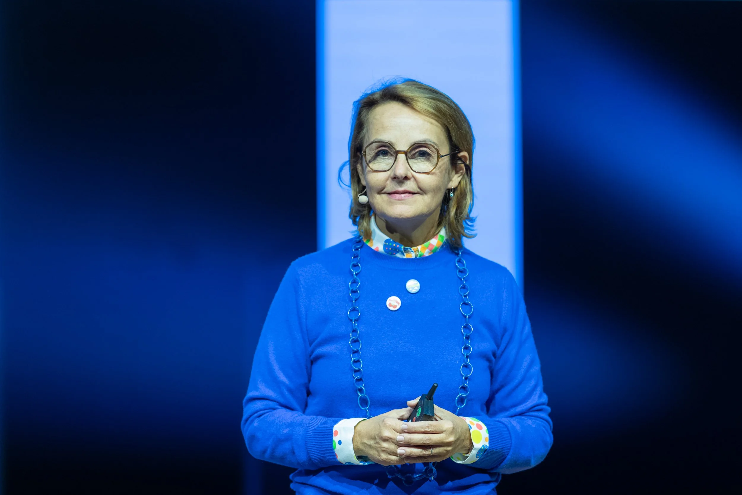 Une femme portant une tenue bleue, des lunettes rondes, des boutons avec des dessins, et un collier en chaîne, se tenant devant un fond sombre avec une lumière derrière elle.