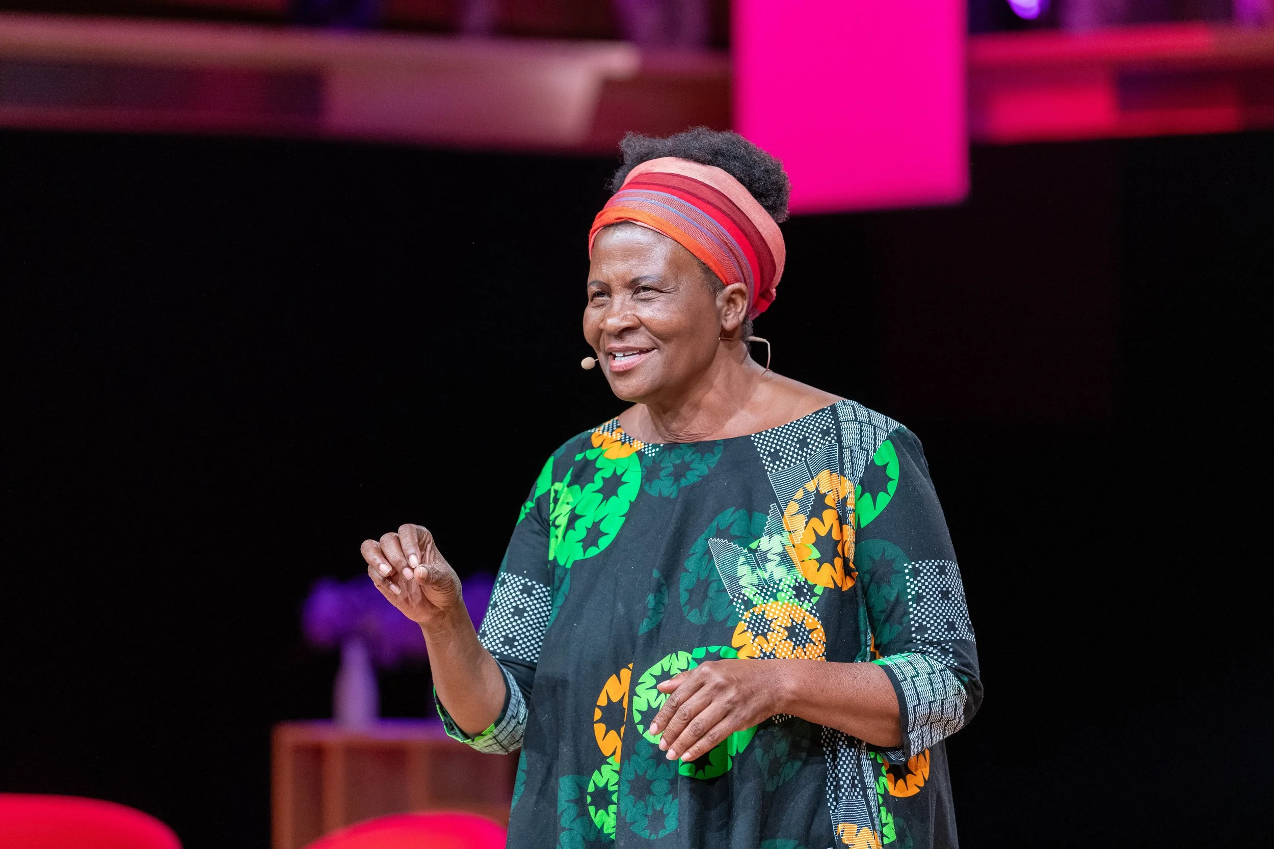 Femme souriante portant un foulard coloré et une robe aux motifs africains, parlant lors d'une conférence sur scène.