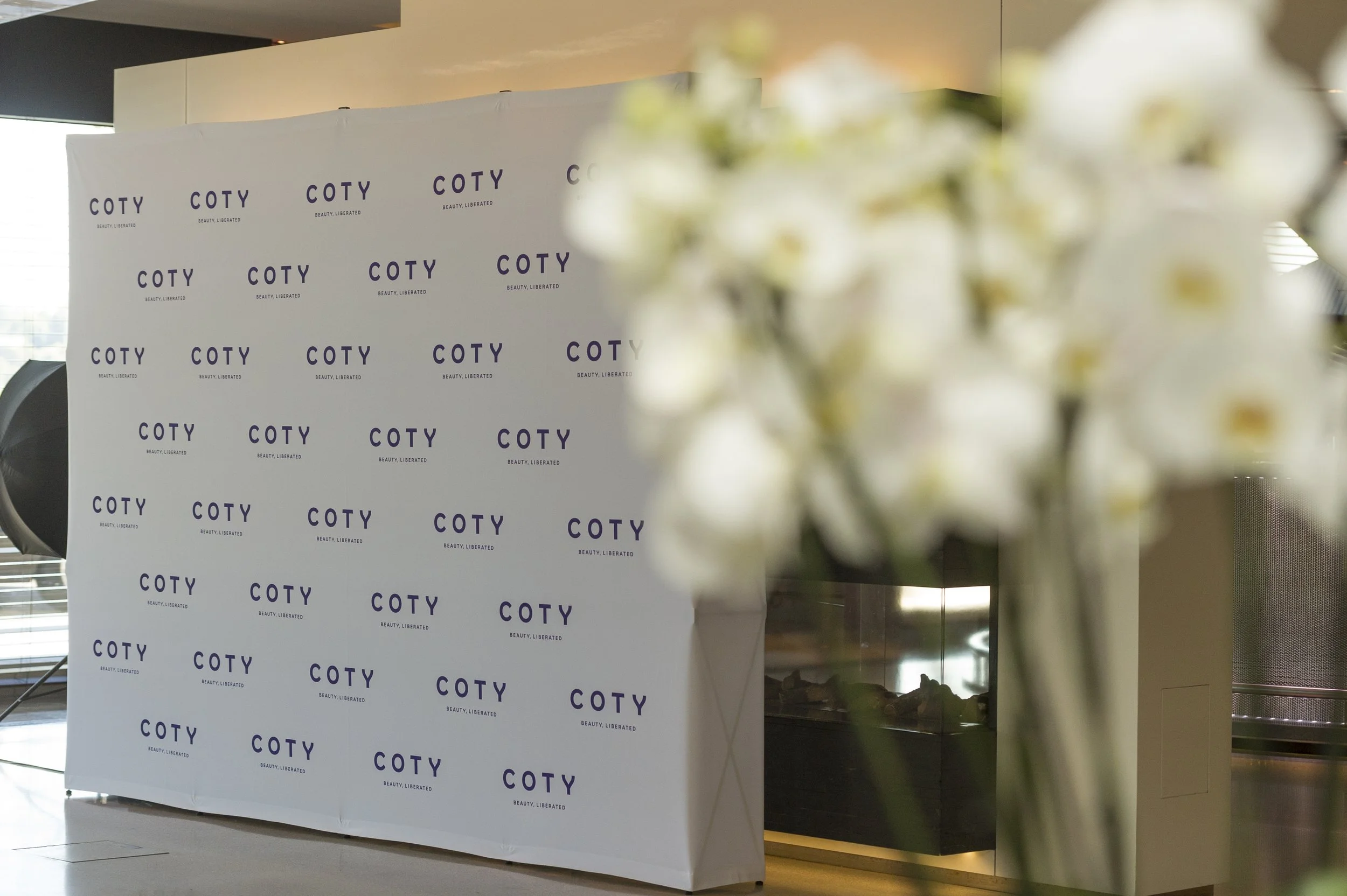 Une toile de fond blanche avec le logo COTY et la phrase 'BEAUTY, LIBERATED' imprimés en violet, située à un événement ou une présentation, avec un bouquet de fleurs blanches floues en premier plan à droite.