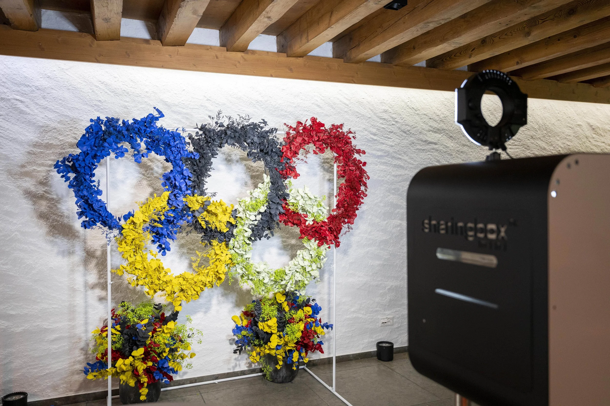 Une installation artistique de fleurs colorées formant les anneaux Olympiques avec des fleurs jaunes, bleues, noires, rouges et vertes, sous un plafond en bois, avec un appareil photo en premier plan.