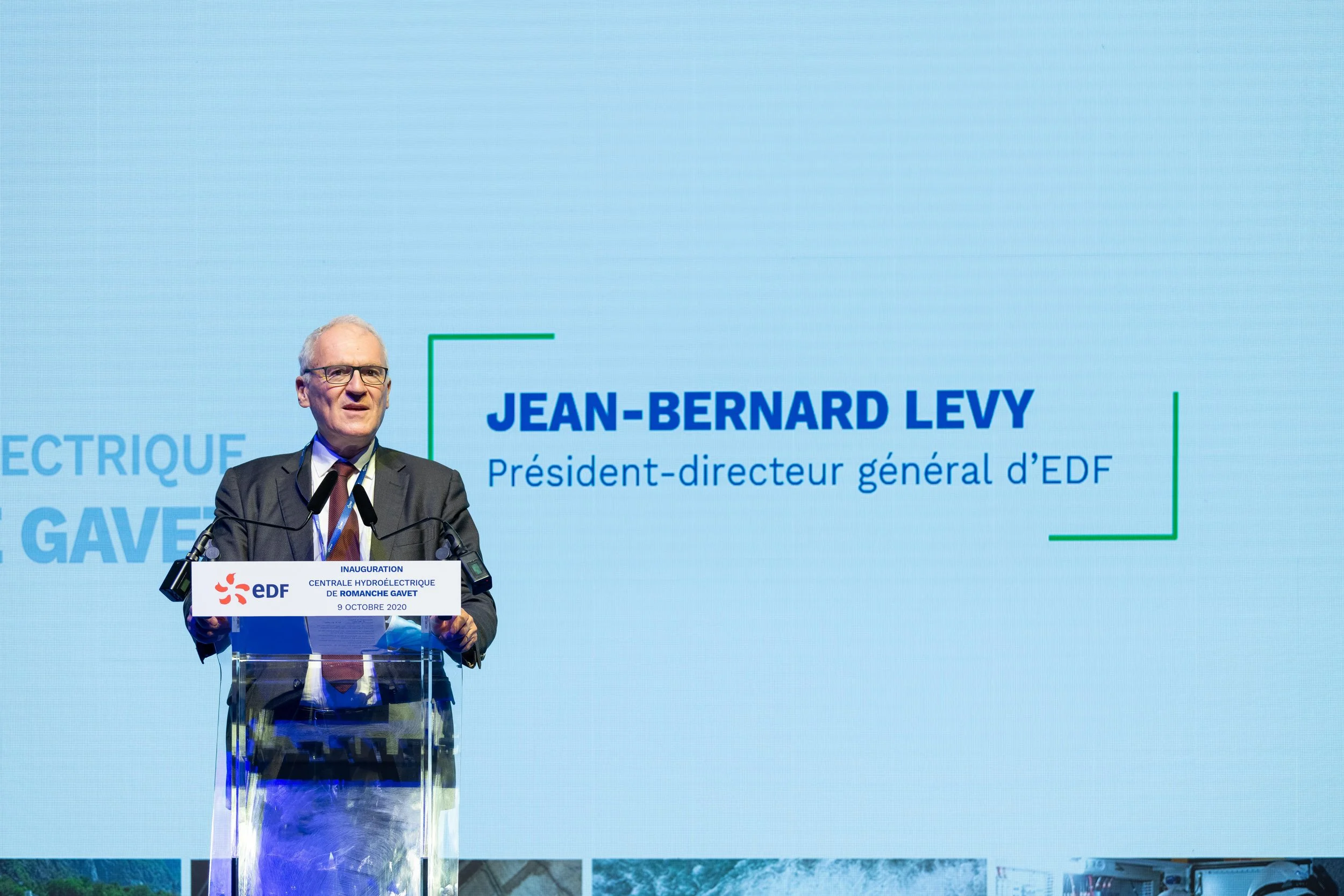 Jean-Bernard Levy, président-directeur général d'EDF, parle lors de l'inauguration de la centrale hydroélectrique de Romanche Gavet le 9 octobre 2020, debout derrière un pupitre avec le logo EDF, avec un grand écran derrière lui affichant son nom.