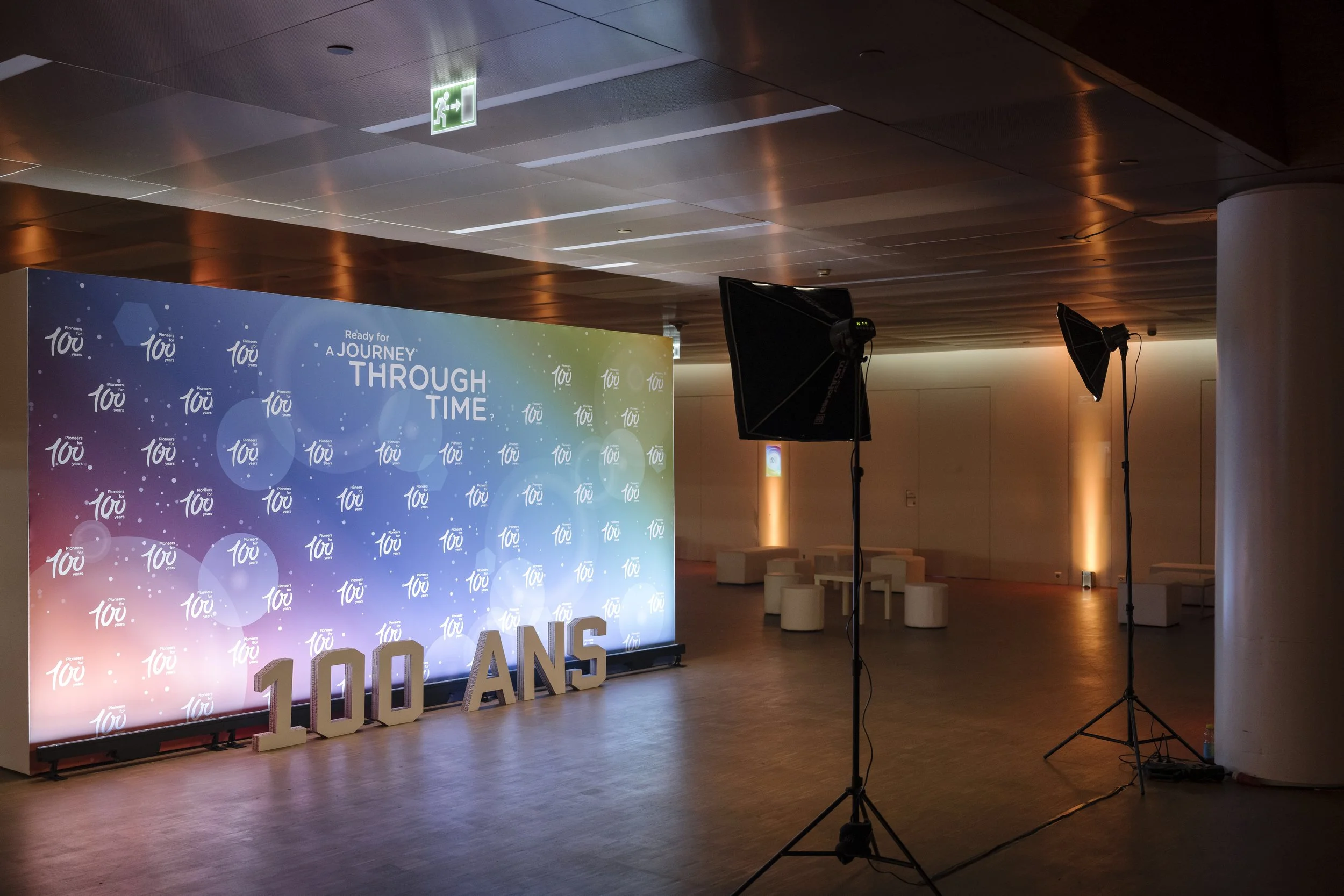 Décoration intérieure pour un événement célébrant le 100e anniversaire, avec un grand panneau lumineux affichant 'Ready for a journey through time' et des lettres indiquant '100 ans', deux spots d'éclairage, et un espace avec des sièges blancs.
