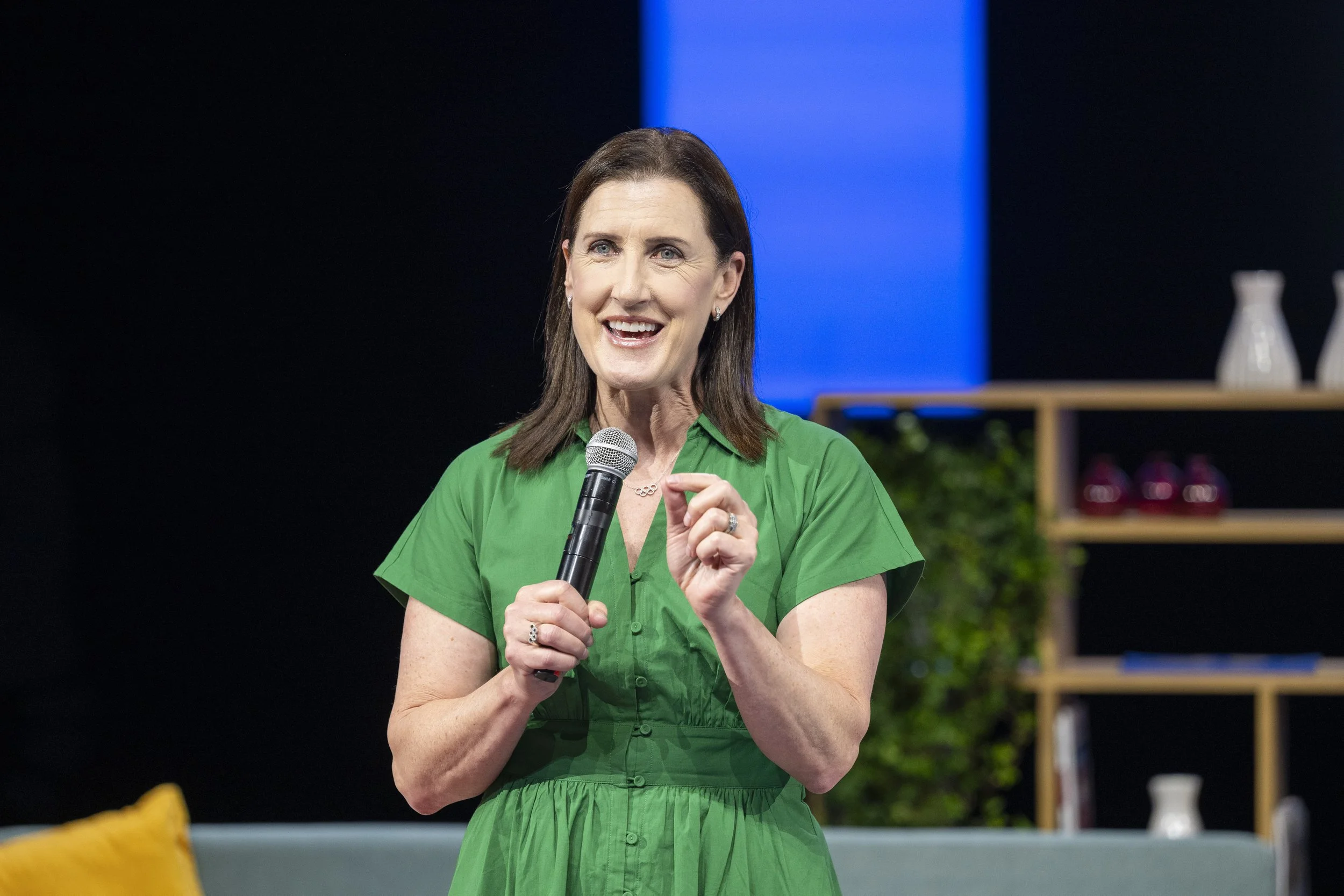 Une femme souriante en robe verte parle dans un microphone sur scène, avec un décor de salon en arrière-plan.