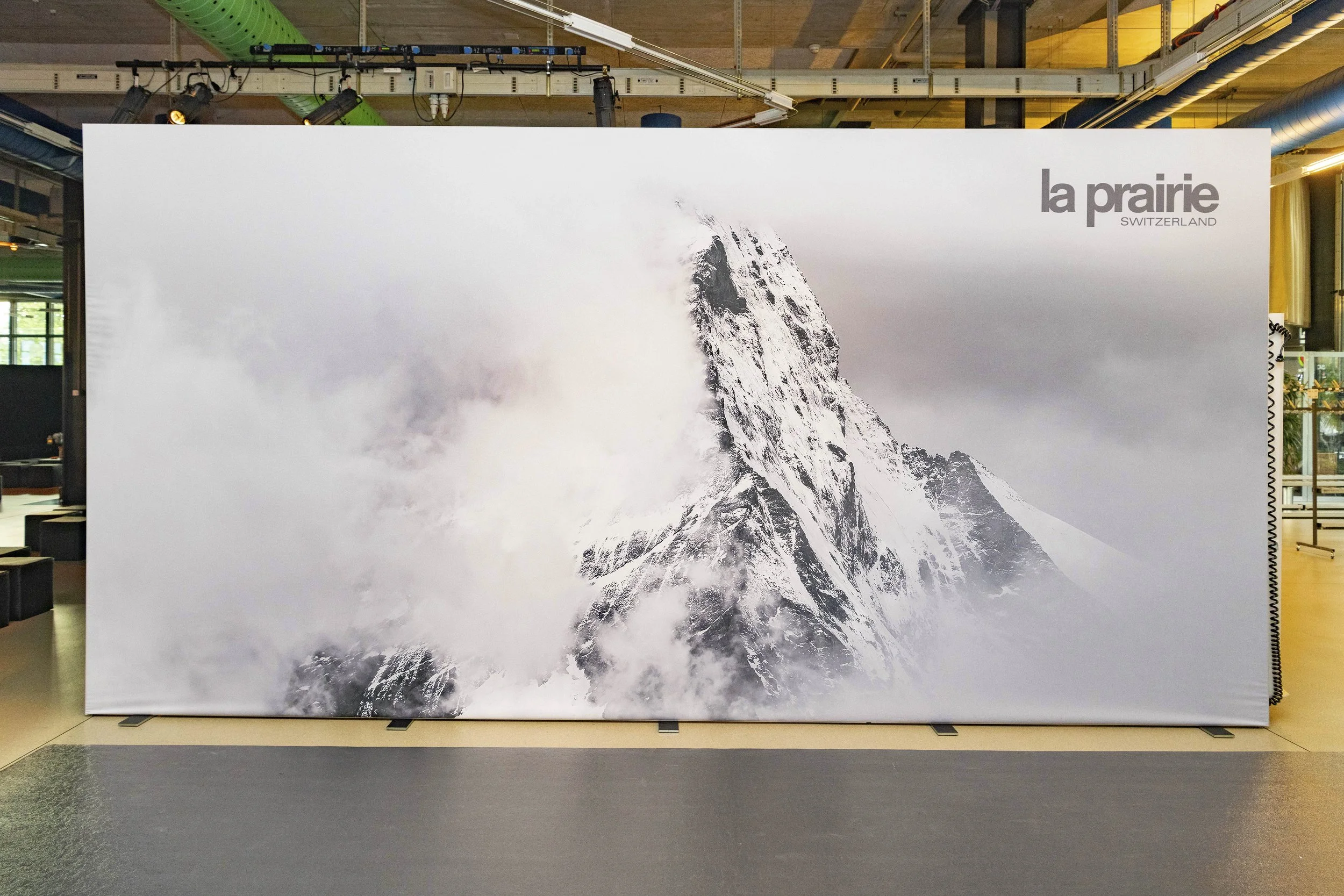 Panneau publicitaire représentant une montagne enneigée, sommet pointu, avec nuages et brouillard, en arrière-plan. En haut à droite, le texte "la prairie" et "SWITZERLAND".