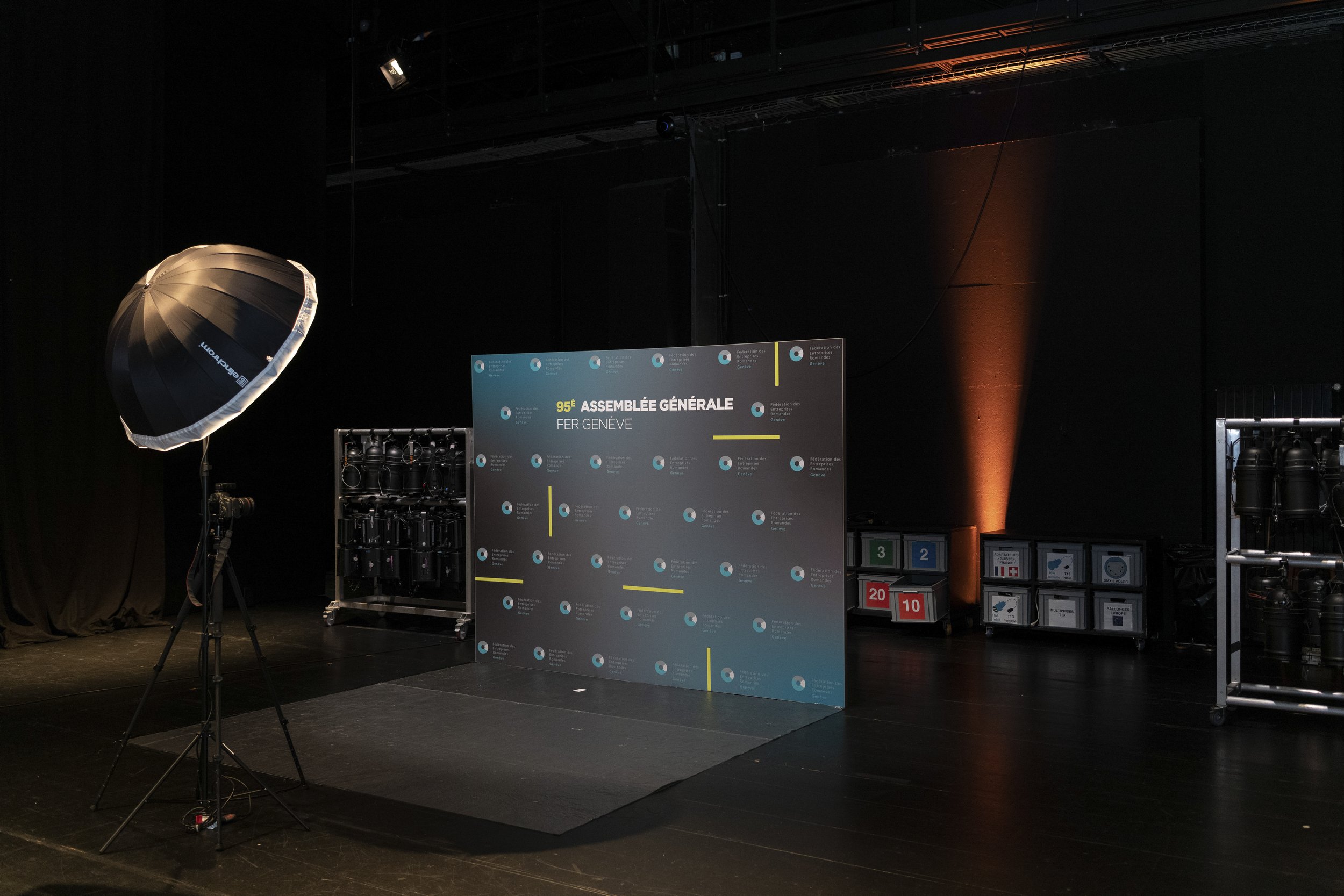 Scène d'une salle de conférence avec un panneau indiquant la 95e Assemblée Générale à Genève. Il y a un éclairage professionnel et des équipements techniques en arrière-plan.
