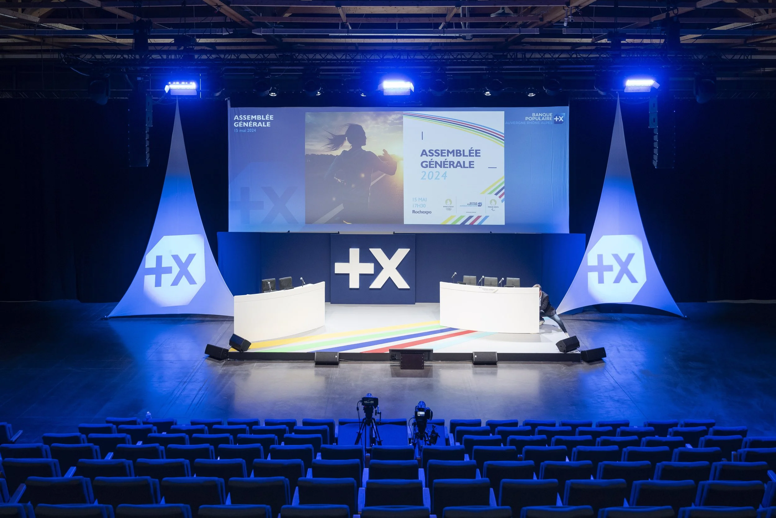Salle de conférence vide avec un grand écran affichant le texte 'Assemblée Générale 2024' et des sièges vides devant la scène.